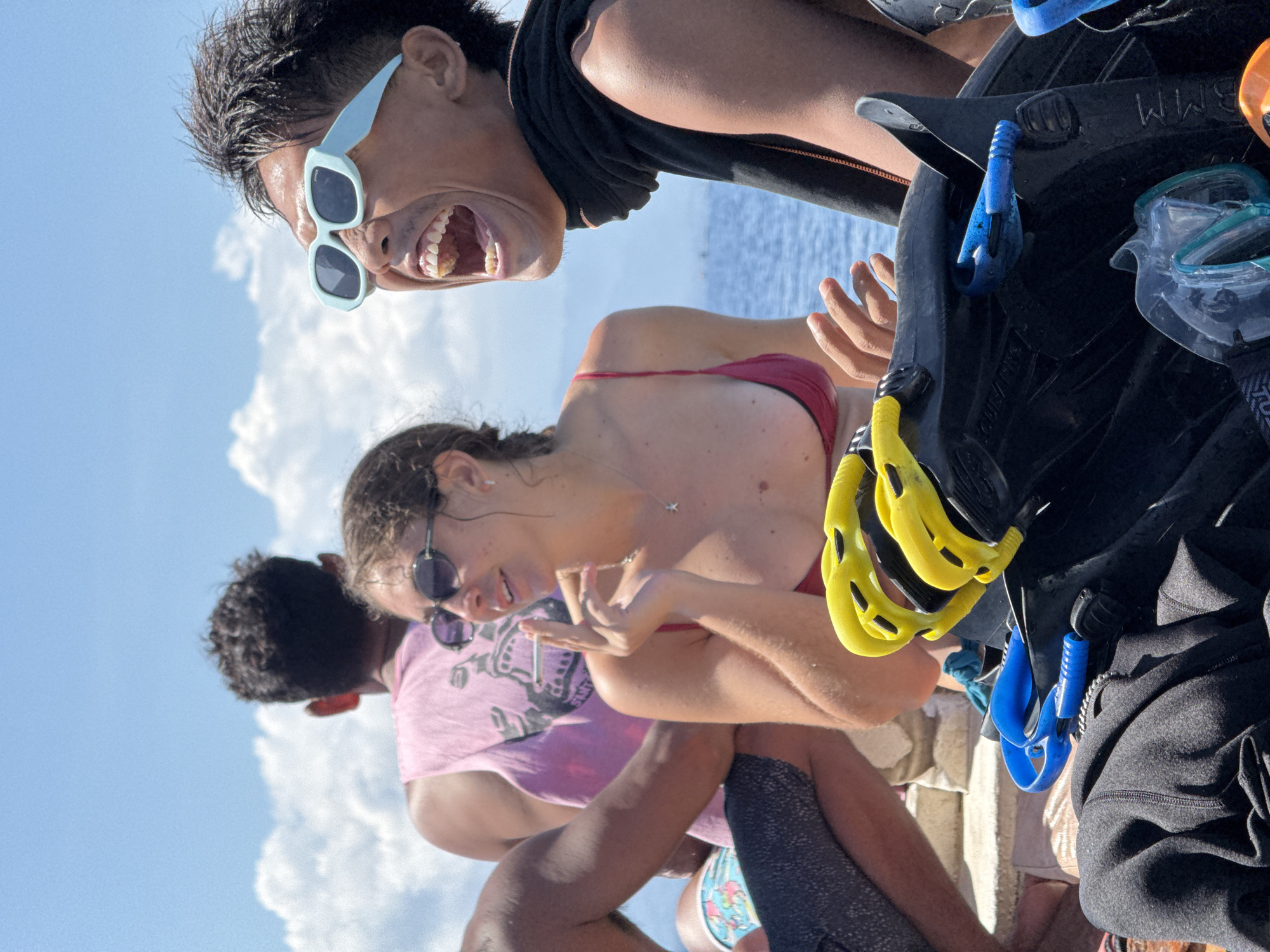 The image shows a group of people on what appears to be a boat in Unknown, Indonesia. The background features a partly cloudy sky and the sea. A man in light blue sunglasses is at the top of the frame, yelling with his mouth open. Below him, a woman in a red bikini and sunglasses is holding a cigarette. A man in a pink shirt is visible to her left. Diving gear, including fins and masks, is in the foreground, obscuring some of the scene.