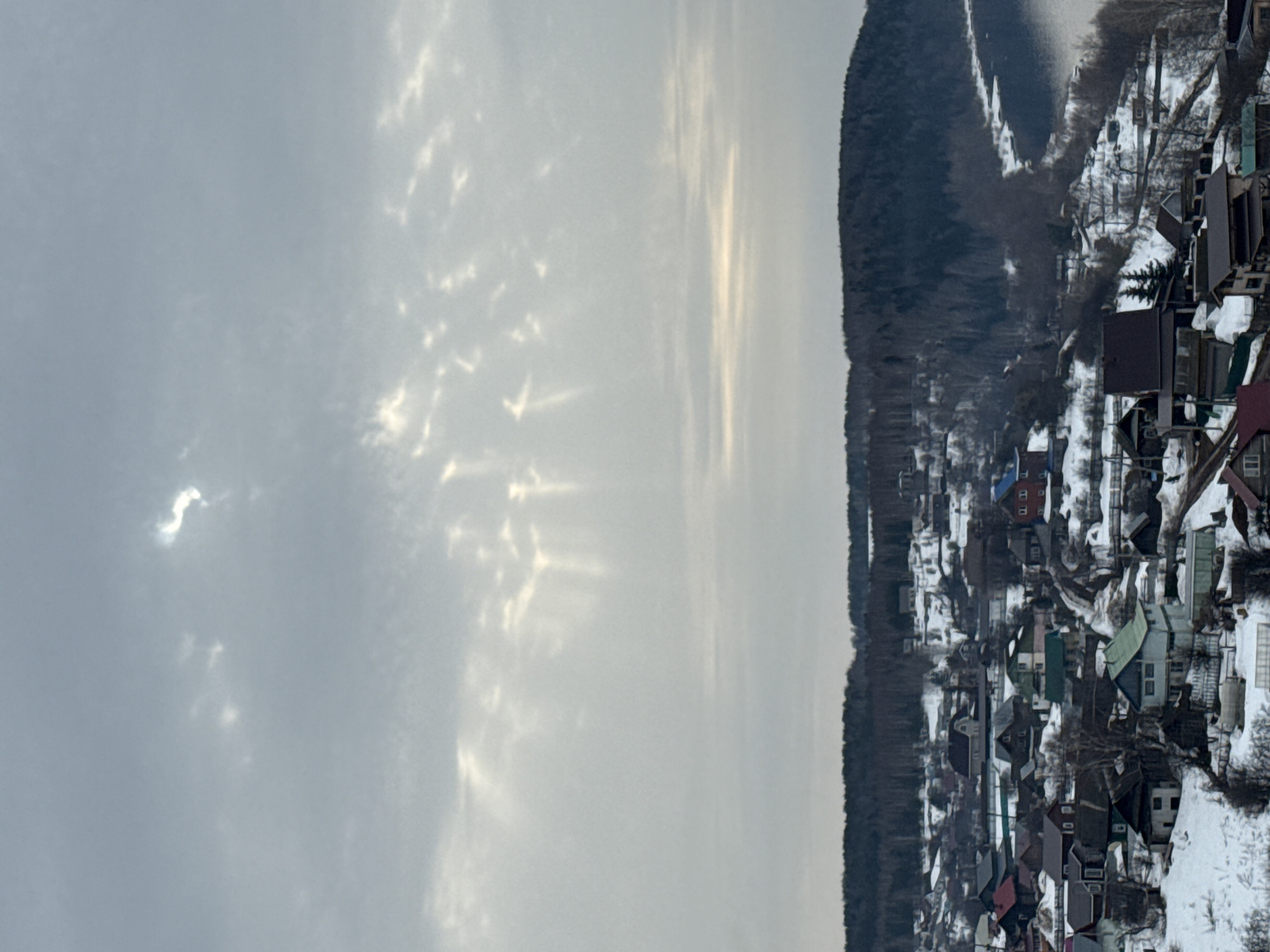 An elevated long shot depicts a winter landscape in Ольховка, Russia, under an overcast sky. The upper two-thirds of the image show a predominantly grey sky with scattered clouds. Sunlight penetrates the cloud cover in multiple locations, forming distinct crepuscular rays that descend towards the horizon, particularly noticeable in the upper left and central right portions of the sky. A bright, irregular, S-shaped luminous anomaly is visible in the upper right quadrant of the sky. The lower third of the image frames a snow-covered village situated on undulating terrain. Numerous detached residential houses with varying roof colors (dark grey, brown, green, red, blue) are visible, dispersed across the snow-dusted slopes. Bare deciduous trees and evergreen conifers are interspersed throughout the village and along the slopes. Unpaved roads and pathways cut through the snow between the structures. In the far right, a dark body of water, likely a river or lake, is visible with snow and ice along its immediate banks, bordering a dense, dark forest line that spans the background horizon from left to right. No individuals or active interactions are visible within the scene.