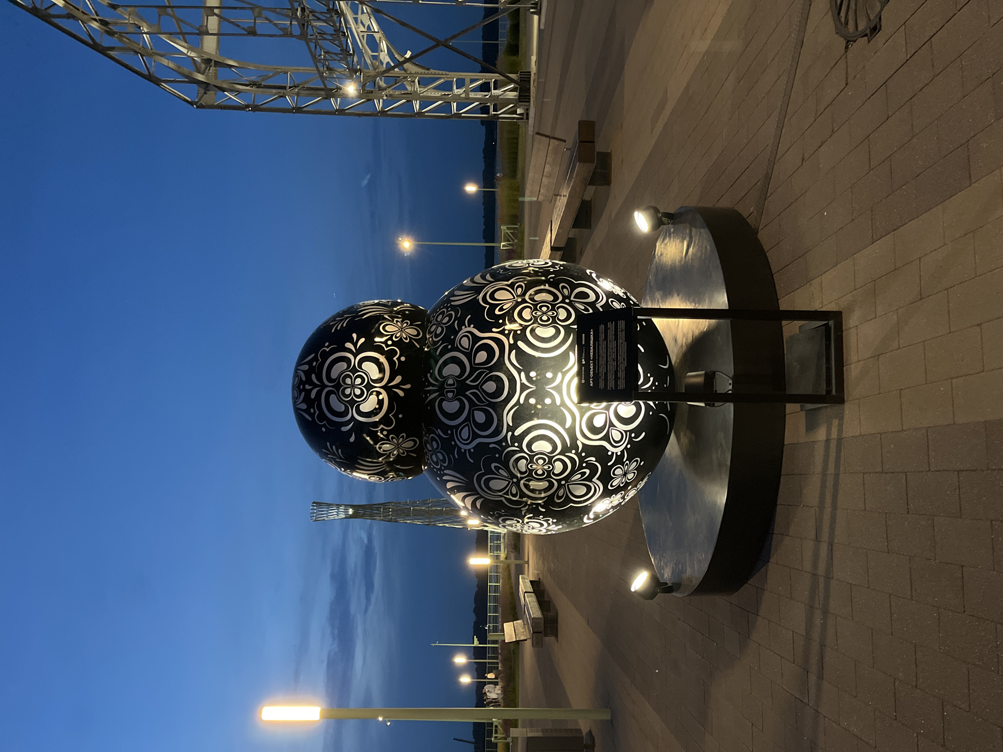 The image shows a sculpture outdoors at dusk. The sculpture consists of two large, black spheres decorated with intricate white floral patterns. It is illuminated by spotlights, casting shadows on the brick-paved ground. A metal frame supports the sculpture, which sits on a circular base. In the background, there are streetlights, benches, and a large, industrial metal structure, possibly part of a bridge or a large building. The sky is a deep blue with some faint clouds, indicating twilight. The overall atmosphere is calm and quiet. There is some text visible on a plaque attached to the sculpture, but it is too blurry to read. The location context provided is Nizhny Novgorod, Russia.
