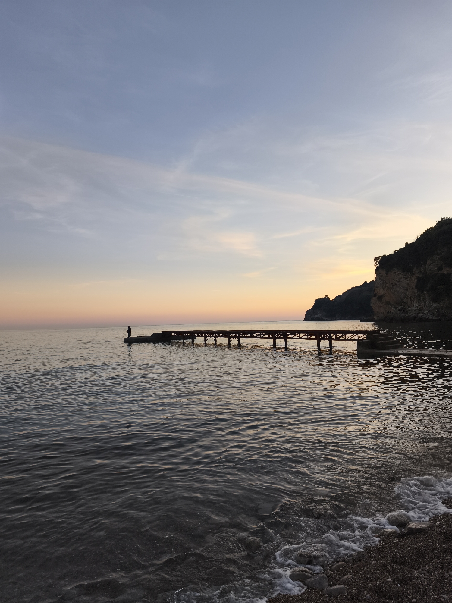 A serene coastal scene unfolds at dusk in an unspecified location. A lone individual stands at the end of a pier that extends into the tranquil water. The pier appears to be constructed of dark wooden planks and supported by beams. The sky transitions from light blue to orange, with a few scattered clouds. The water is gently rippling, reflecting the soft colors of the sunset. A rocky cliff covered with trees is visible on the right side of the frame, marking the boundary of a small cove. Waves are lapping gently onto a pebble beach in the foreground. The atmosphere is calm and peaceful.