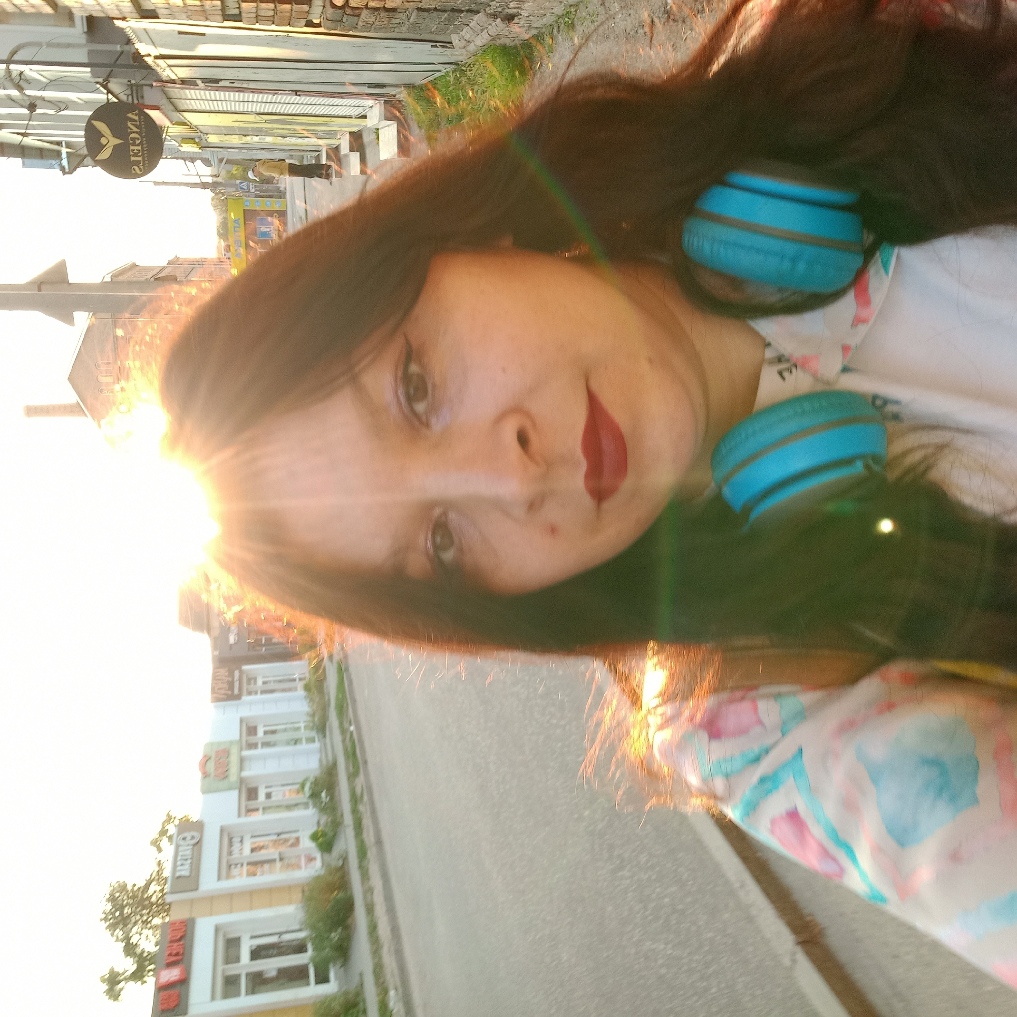 The image is a selfie taken from the perspective of a person walking on a sidewalk in Kropyvnytskyi, Ukraine. The sun is shining brightly, creating lens flares and glare that obscure parts of the scene, particularly the upper portion of the image.
In the foreground, the face of a woman is visible. She has dark hair, makeup including red lipstick, and is wearing blue headphones around her neck. She is also wearing a light-colored shirt with a colorful abstract pattern.
The background shows a street scene with buildings lining one side. On the left, there are several storefronts with signs that appear to be in Ukrainian or Russian, including one that reads "BAZTAR". On the right, there is a building with brickwork and a circular sign that reads "ANGELS" in reverse, indicating it's seen from a reflection or a reversed image. A person in a yellow jacket and a white cap is visible standing near the storefronts on the right. The road is paved and appears to be an urban street. Greenery, likely grass and bushes, is present along the side of the road and in front of the buildings.