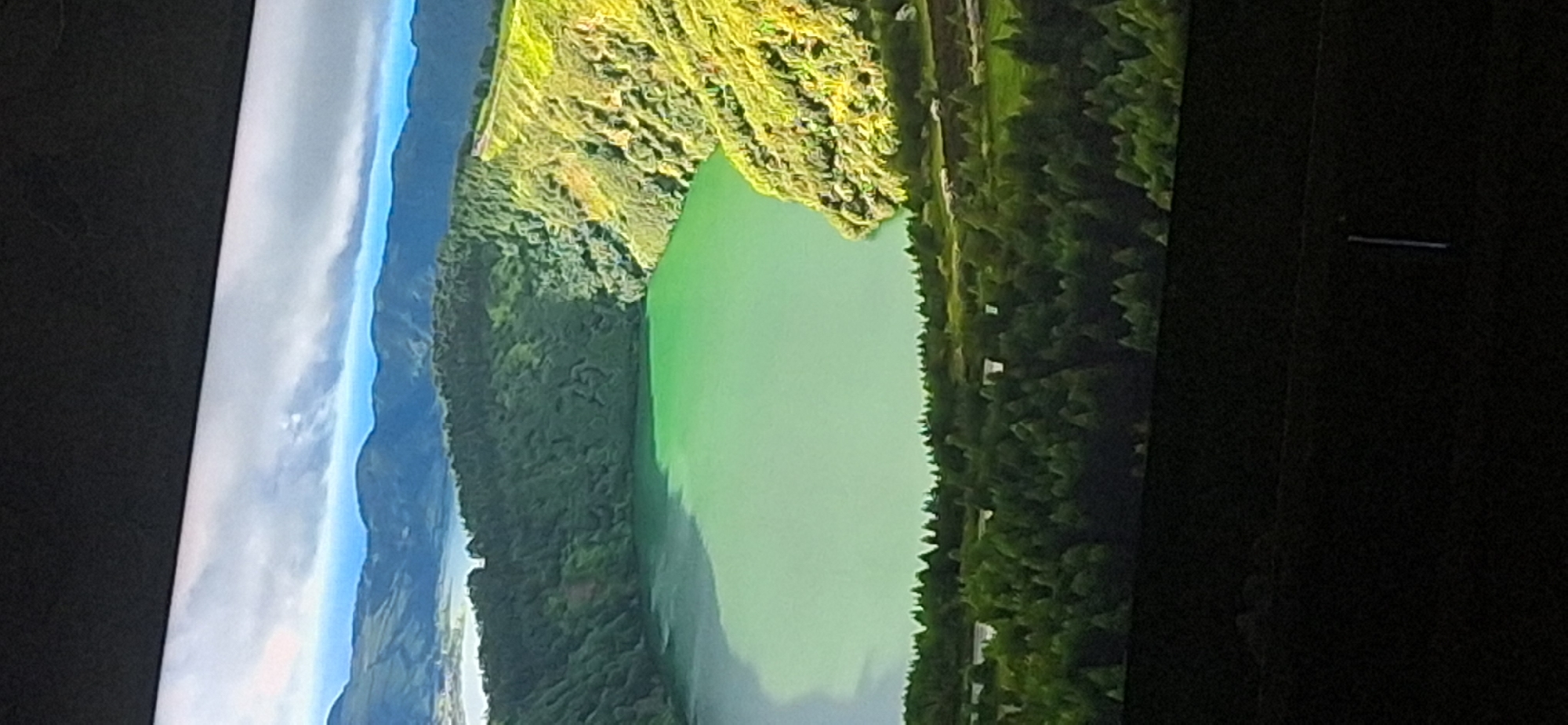 The image depicts a panoramic view of a volcanic crater lake, with lush green vegetation covering the crater walls and surrounding hills. The lake itself has a striking emerald-green color. In the background, a range of mountains can be seen, with a hazy sky above. The foreground shows a dense forest. There is no indication of the city of Bila Tserkva, Ukraine, in this image.