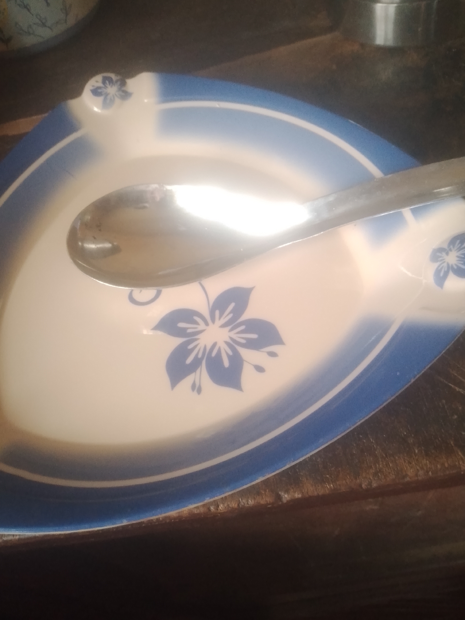 A spoon rests on a white plate with a blue floral pattern. The plate has a blue rim and appears to be made of ceramic. The spoon is made of metal and has a curved shape, typical of a soup spoon. The background is a dark, wooden surface, possibly a table. In the upper left corner, a portion of another object, possibly a bowl, is visible. The lighting suggests an indoor setting, and the overall scene is one of domestic simplicity. There are no people visible in the image, and no text is present. The setting does not provide any specific clues about the time of day, weather, or emotional state. The location context of Jauro Yinu, Nigeria, is not visually represented in the image itself.