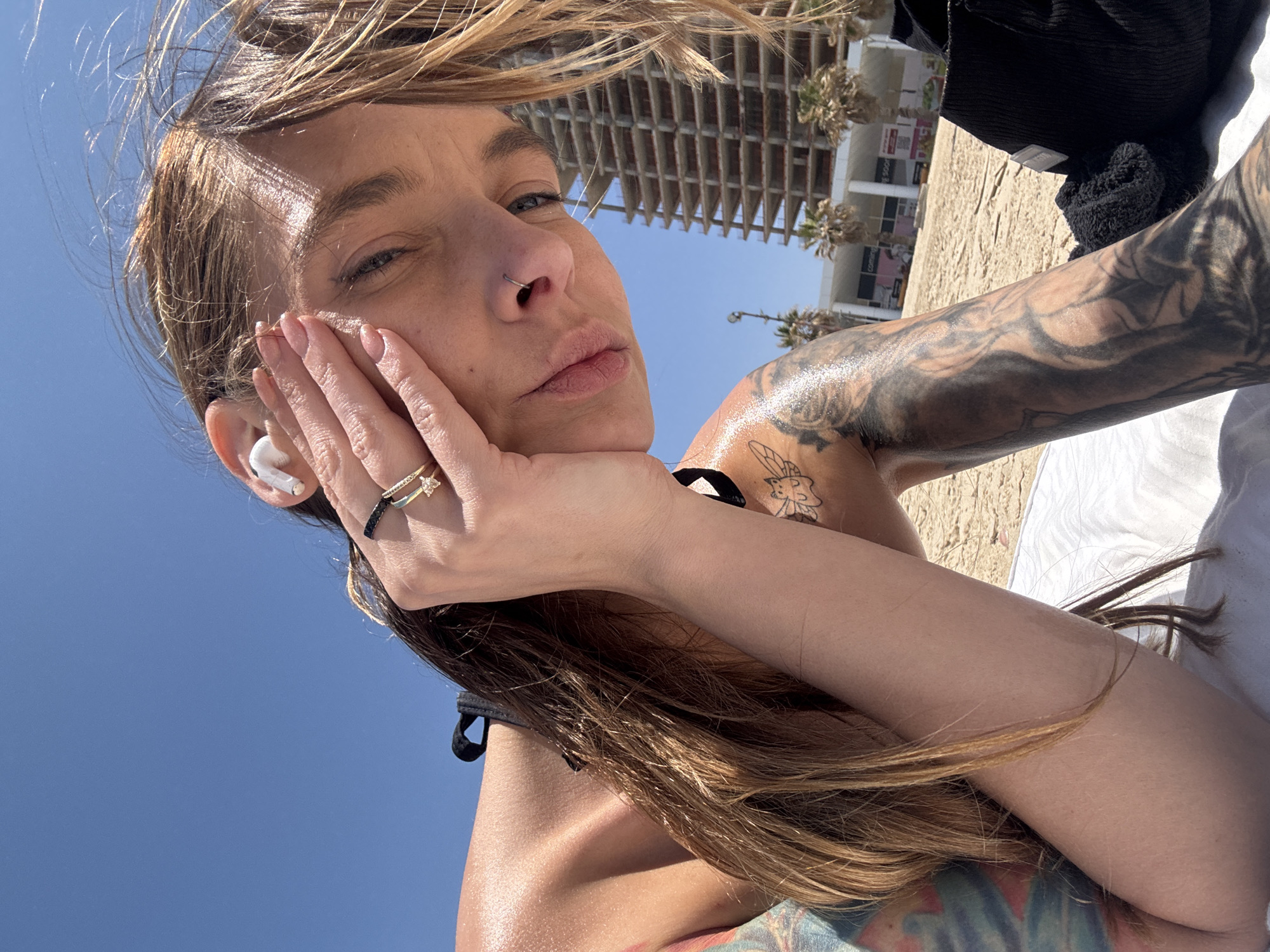 A selfie shows a woman with light brown hair, a nose ring, and visible tattoos on her right arm and shoulder. She is wearing rings on her left hand, which is supporting her head. An earbud is visible in her left ear. A building with multiple floors is visible in the background, along with palm trees. The image was taken on a sunny day in Bat Yam, Israel. The background shows a beach with sand. She is wearing a black bikini top.