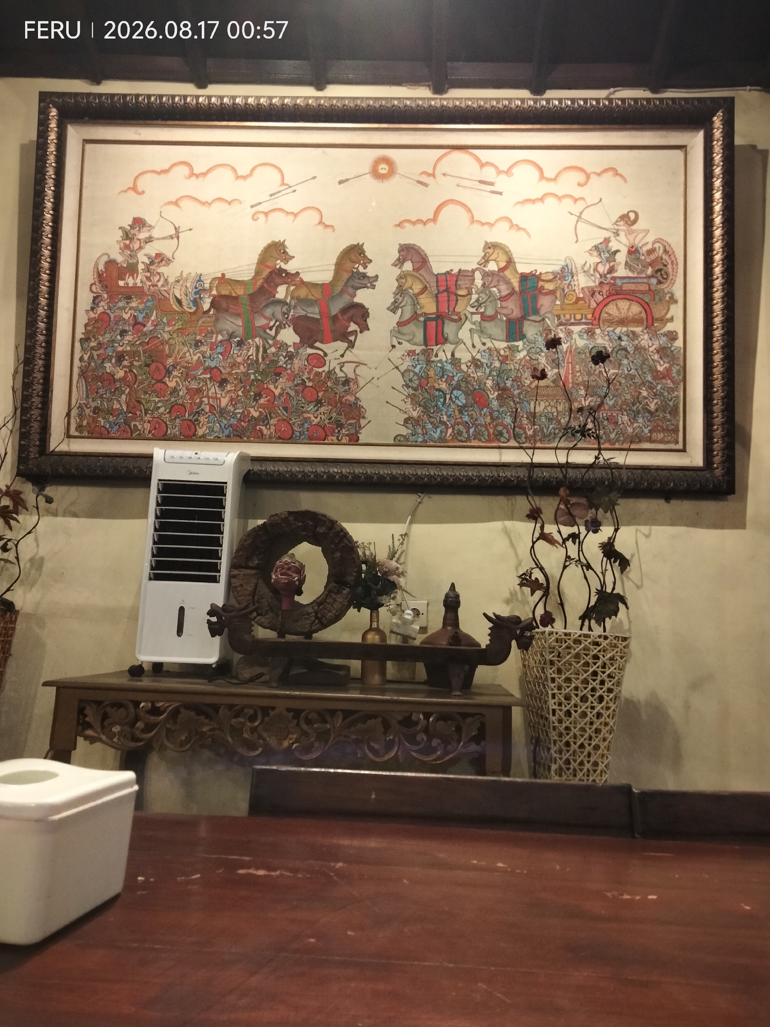 The image depicts a still life composition indoors, likely a dining or living area. Dominating the background is a large framed painting featuring a chaotic battle scene with chariots, horses, and numerous figures, rendered in a vibrant, traditional style. In the mid-ground, a white evaporative cooler stands to the left of center. To its right, on a carved wooden table, rests a circular wooden artifact with a red-faced figure inside. Further to the right, a woven basket contains a decorative arrangement of dried branches and leaves. The foreground shows a dark wooden table with a white rectangular object on its left side. The date stamp in the upper left corner reads "FERU | 2026.08.17 00:57", suggesting the image was captured in Sukodono, Indonesia.