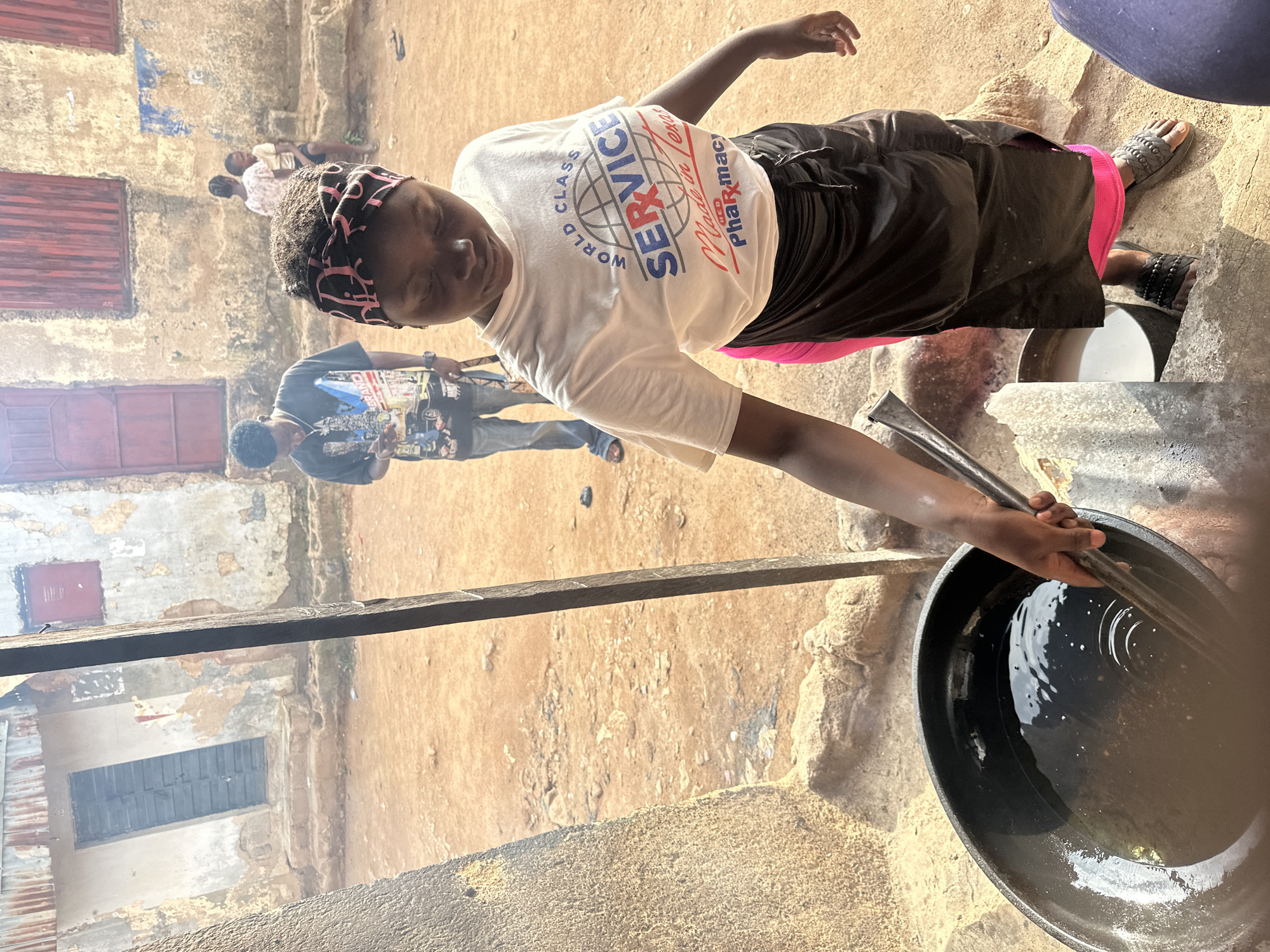 The image depicts an outdoor scene in what appears to be a developing urban environment. The foreground is dominated by a young person wearing a white t-shirt with a "SERVICE" logo and a black skirt over pink fabric. They are holding a metal utensil and appear to be interacting with a large black pot filled with dark liquid. To the left, a textured stone structure is visible. In the background, two other individuals are present: a male figure in a graphic t-shirt and jeans looking at his phone, and two children sitting. The environment features weathered buildings with peeling paint and corrugated metal roofing, set against a dusty, unpaved ground. The overall impression is of a casual, everyday activity taking place in a modest setting.