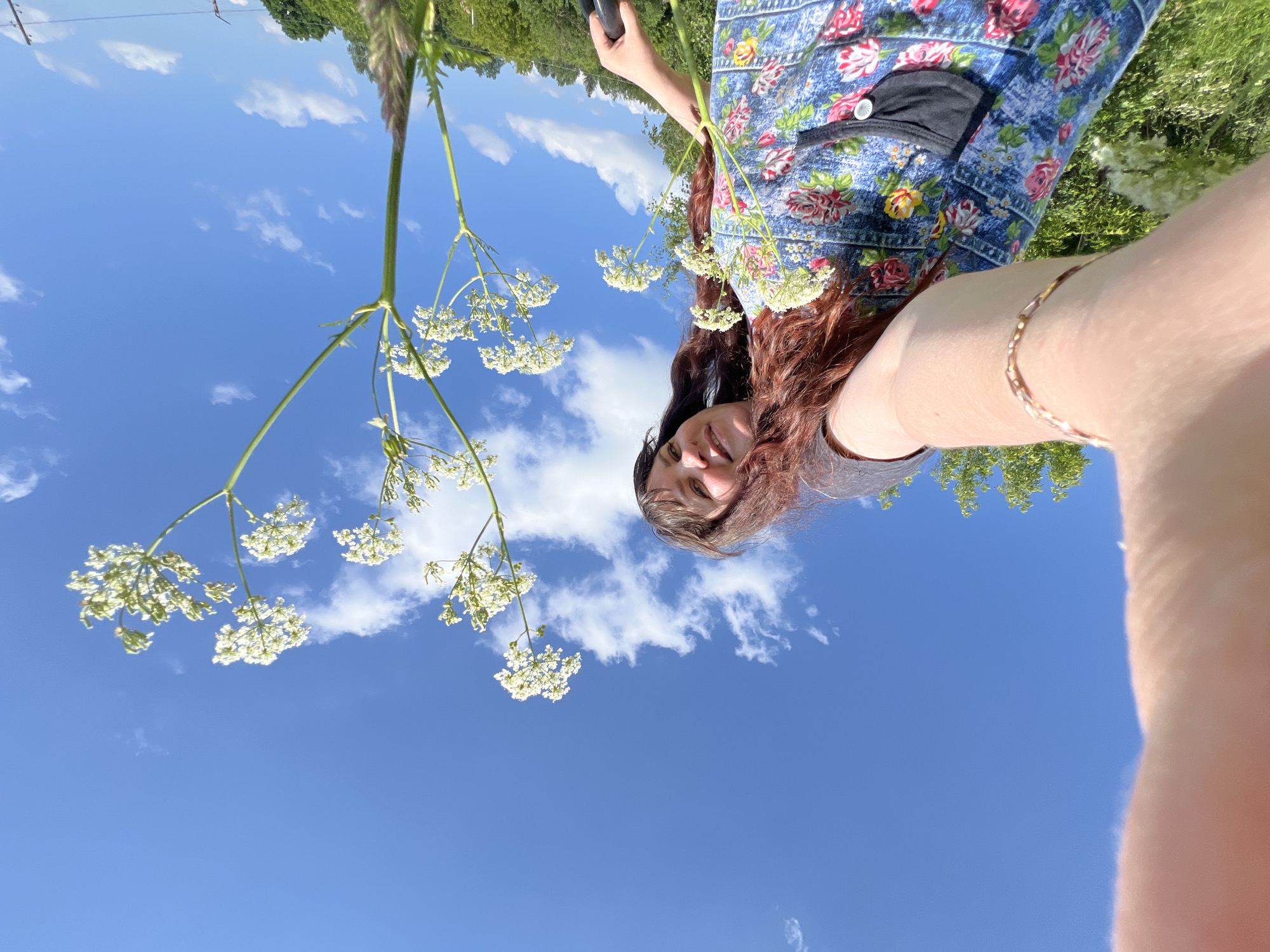 A low-angle, wide shot captures a young woman taking a selfie in a field under a bright blue sky with scattered white clouds. The woman, with long, dark, wavy hair, is wearing a denim apron with a floral print of pink roses and green leaves. She has a light skin tone and is smiling at the camera. Her right arm is extended towards the camera, and a bracelet with small, colorful beads adorns her wrist. In the foreground, several tall, delicate white wildflowers with umbel-shaped clusters of tiny flowers are prominent, reaching towards the sky. The background shows a slightly blurred green landscape with trees and foliage, suggesting an outdoor, rural or semi-rural setting. The presence of electric wires visible in the far background indicates proximity to infrastructure, but the overall impression is natural and serene. The specific location, Panteleevka, Russia, is noted.
