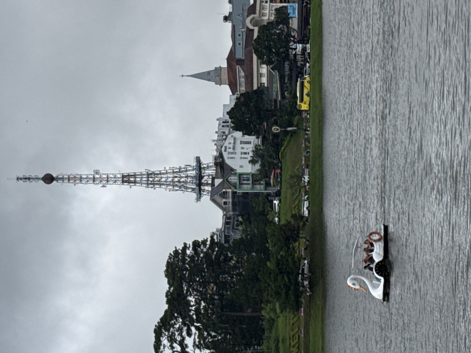 This image depicts a serene lakeside scene in Da Lat, Vietnam, under an overcast sky. In the foreground, a white swan-shaped pedal boat, carrying at least two people, glides on the lightly rippled surface of the lake. One person, wearing a light-colored top, is clearly visible operating the pedals.
The left bank of the lake features a verdant strip of grass, behind which a road is lined with various parked vehicles, including a distinctive yellow mini-bus. Beyond the road, the city's architecture is visible, dominated by a tall, lattice-structured tower on the left, reminiscent of the Eiffel Tower, complete with a spherical element near its top. Further along the skyline, a building with a prominent pointed steeple or spire stands out. Numerous other buildings, featuring diverse architectural styles and colors, are nestled among abundant green trees, which also line the lake's edge. The sky is a uniform light grey, indicating cloudy weather. No visible text is legible in the image.