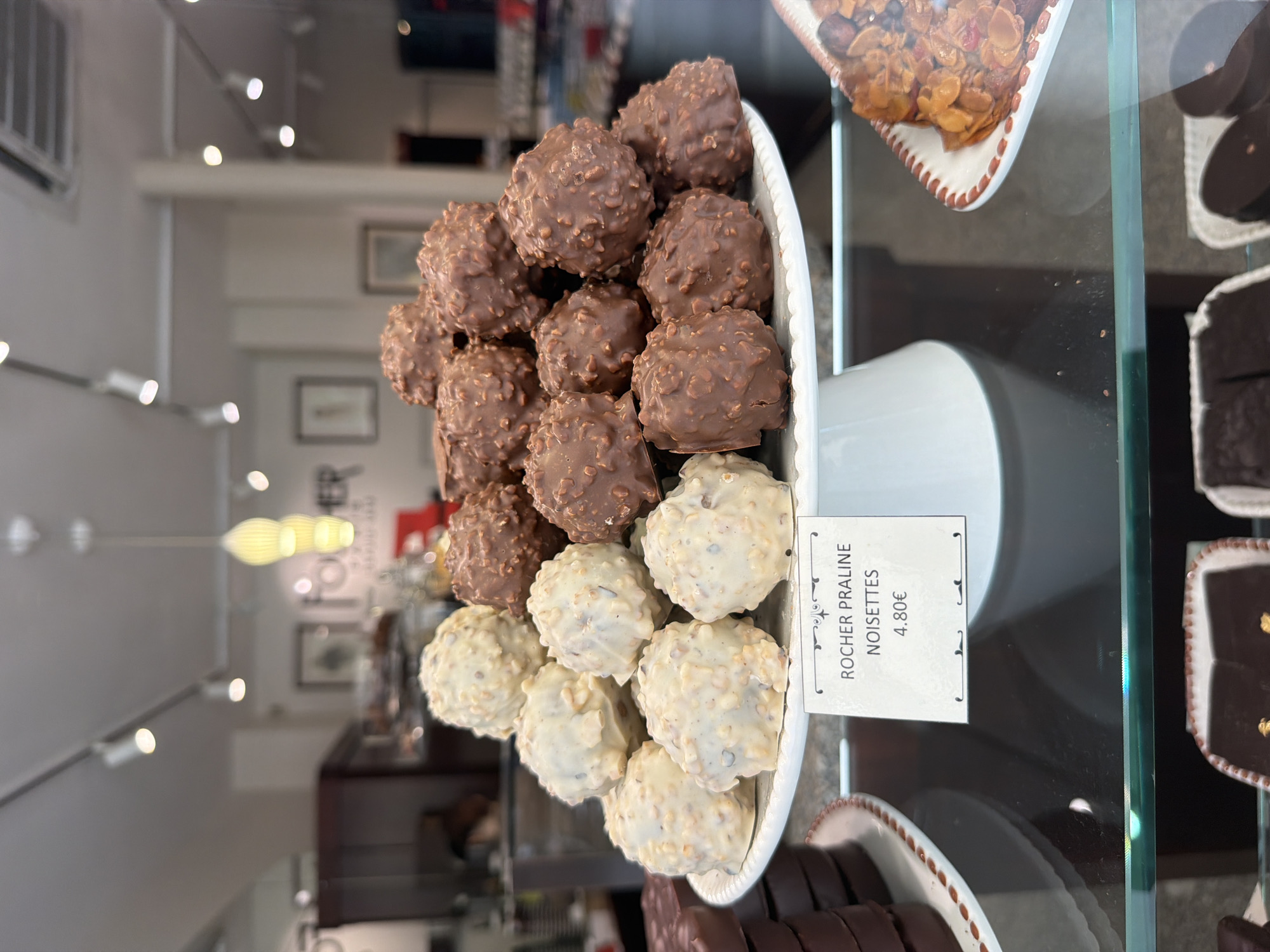 The image shows a display case of chocolates in a store, likely in Paris, France. The main focus is a two-tiered, light grey display stand holding chocolates coated in nuts. The top tier holds dark chocolate or milk chocolate balls covered in nuts, while the bottom tier has white chocolate balls covered in nuts. A sign is visible that reads "ROCHER PRALINE NOISETTES 4.80€". Other sweets are visible in the periphery of the display case, including chocolate discs with golden accents, caramel candies, and squares of what appears to be dark chocolate fudge. The store seems well-lit with recessed lighting and light coming through the windows. Through the glass, in the background, there is a blurred view of the interior of the store, including framed pictures on the wall and some hanging lights.
