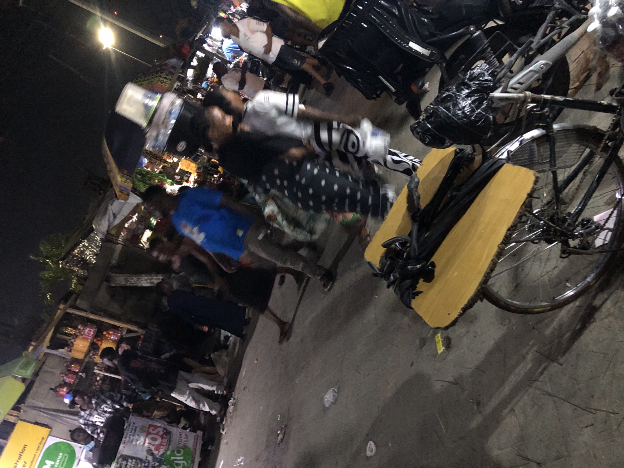 This is a street scene in Moba, Nigeria at night. A partially visible bicycle is in the foreground on the right, resting on a wooden plank with black objects bundled on top. A blurry group of people is in the middle ground walking left to right. The background shows what appears to be a market or commercial area with lit stalls and crowds of people.