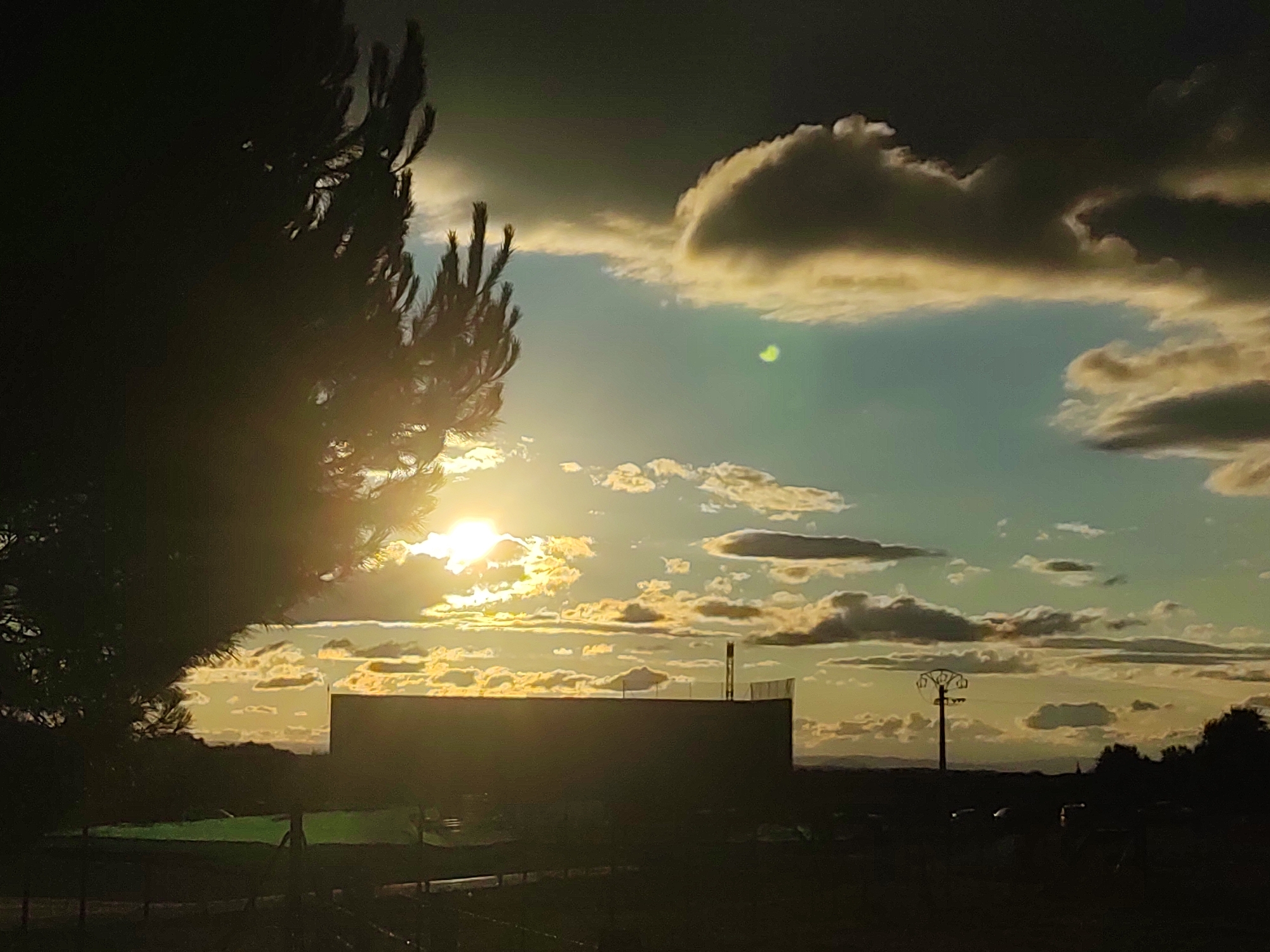 This image captures a sunset scene in Sobradillo, Spain, with dramatic clouds and a silhouetted landscape. In the foreground, a dark tree on the left partially obscures the bright sun, which is low in the sky and casting a golden glow. A large, dark, rectangular structure dominates the mid-ground, its top edge silhouetted against the sky. A tall, dark pole with a cross-shaped top is visible on the right side of this structure. Further in the distance, the horizon is lined with dark trees and hills. The sky is a mix of dark, heavy clouds at the top and lighter, scattered clouds illuminated by the setting sun, creating a dynamic contrast of light and shadow. The ground in the foreground appears to be a dark, possibly paved area with some faint outlines of objects, and a patch of green grass is visible to the left of the large structure.