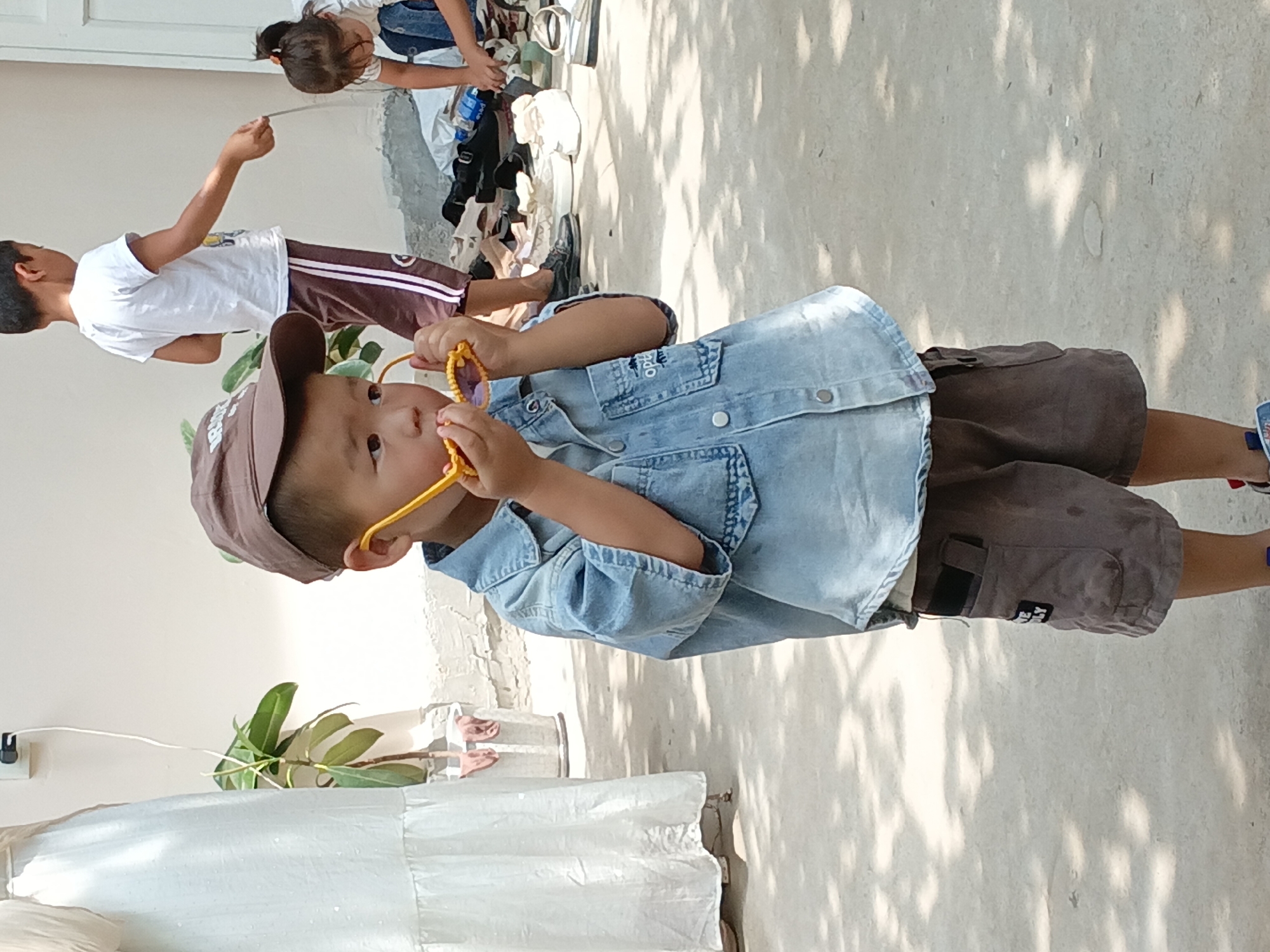 This image, taken in Osh, Kyrgyzstan, depicts a young boy wearing a brown baseball cap and a denim shirt. He is holding a pair of yellow sunglasses up to his face, seemingly examining them. The boy is also wearing brown shorts. In the background, another child, a boy, is wearing a white t-shirt and dark shorts with white stripes, and is interacting with a pile of shoes and bags. Further in the background, a young girl in a white shirt and blue jeans is also engaged with the shoes. A person in a long white dress stands to the far left of the frame. The ground is a concrete surface with dappled sunlight and shadows from overhead foliage. The overall setting appears to be an outdoor area, possibly a yard or courtyard, with a wall in the background.
