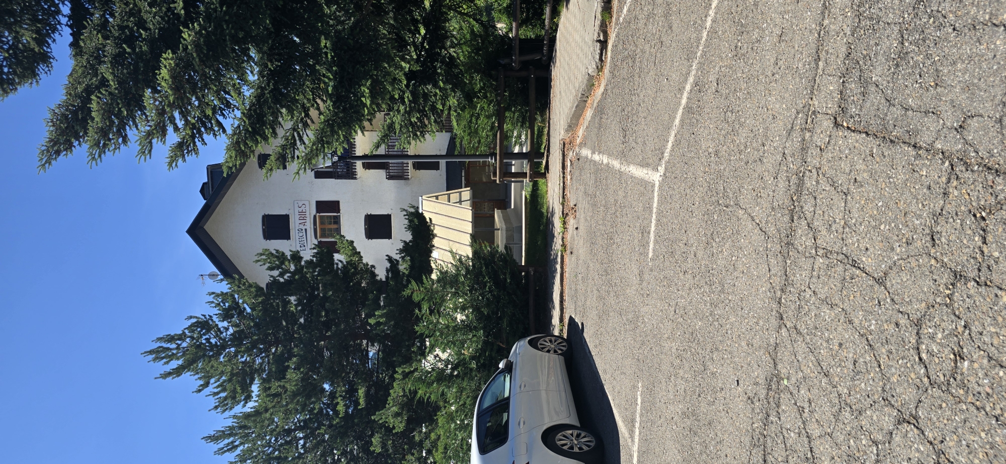 In Sallent de Gállego, Spain, a white hatchback car is parked in a cracked asphalt parking lot. Behind the car, a white building with a dark roof, identified by the signage "EDIFICIO ARIES", stands partially obscured by tall green pine trees. The building features multiple windows with dark shutters and a small balcony. In front of the building, a light-colored, angled canopy covers an entrance area. A lamppost is positioned near the building's entrance. The scene is set under a clear blue sky on a sunny day.
