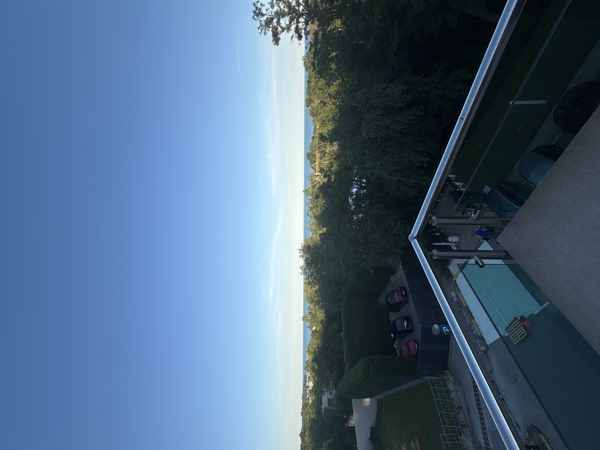The image depicts a clear blue sky above a distant horizon where a body of water meets the land. Below the sky, lush green trees form a dense canopy, with hints of buildings nestled within the foliage. In the foreground, a balcony railing with a metallic finish overlooks a residential area. Several cars are parked on a paved surface. A small, empty swimming pool is visible, with a pallet and an orange traffic cone on its edge. A person in a blue shirt is standing near the pool. The overall scene suggests a tranquil, possibly suburban or resort-like environment during daylight hours.