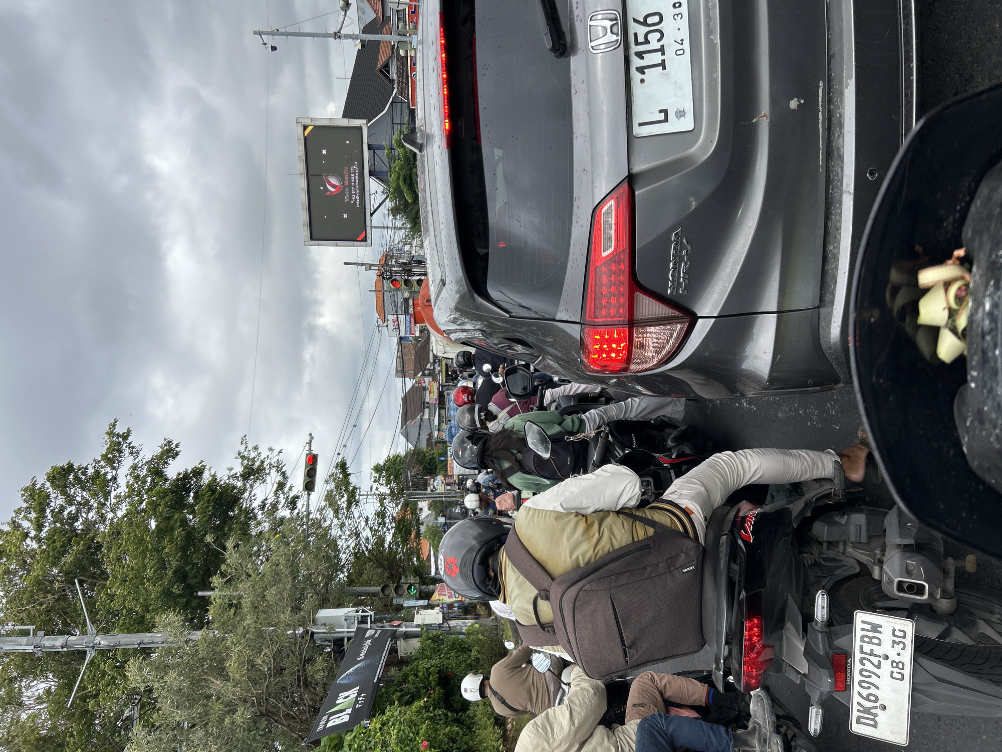 The image depicts a congested street scene in Denpasar, Indonesia, likely during daylight hours under overcast skies. The primary focus is on a traffic jam involving numerous motorcycles and a few cars. In the foreground, a black motorcycle is visible, with a rider wearing a helmet and a backpack. To the right, a grey Honda HR-V is stopped, its taillights illuminated. The license plate reads "L 1156". Further ahead, a dense flow of motorcycles fills the road, with riders in various attire and helmets, indicating a typical urban traffic situation. The environment includes buildings lining the street, trees, traffic lights, and utility poles. A large digital billboard with a logo and contact information for "Vista Media" is prominently displayed overhead, suggesting the presence of advertising and commerce. The overall impression is one of a busy urban intersection experiencing significant traffic volume.