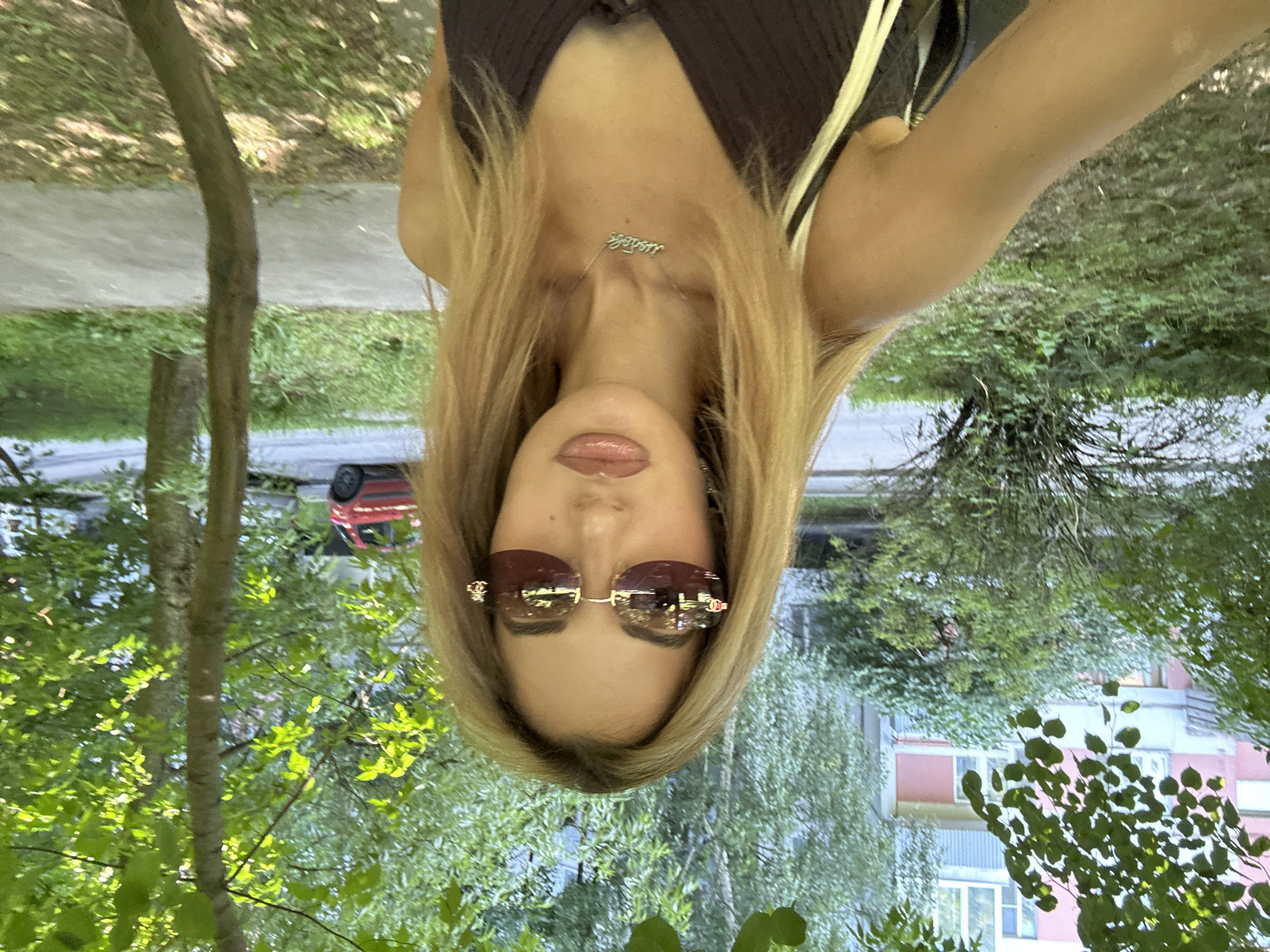 A woman with blonde hair and sunglasses stands outdoors, a building with pink and white siding visible in the background. She is wearing a dark top and a necklace. The scene appears to be a residential area, possibly in Moscow, Russia, with trees and a red car in the distance.