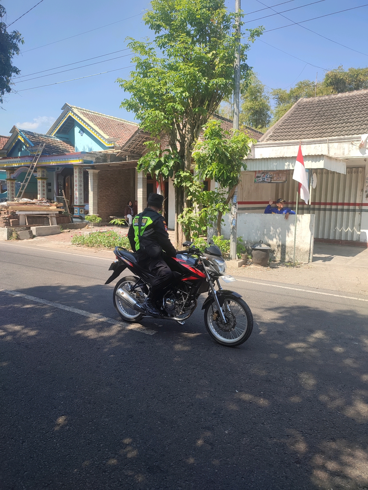 In the city of Nganjuk, Indonesia, under a clear blue sky, a person wearing a black uniform with a bright green reflective vest rides a black and red motorcycle on an asphalt road. The rider is facing away from the camera. Behind the road, several houses with terracotta tiled roofs are visible, some with colorful decorative elements. Trees with lush green foliage line the street. On the right side of the road, an Indonesian flag is displayed on a building. Another person in a blue shirt is standing near the flag. The environment appears to be a residential area with a mix of traditional and modern housing. Shadows from trees are cast across the road and the motorcycle.