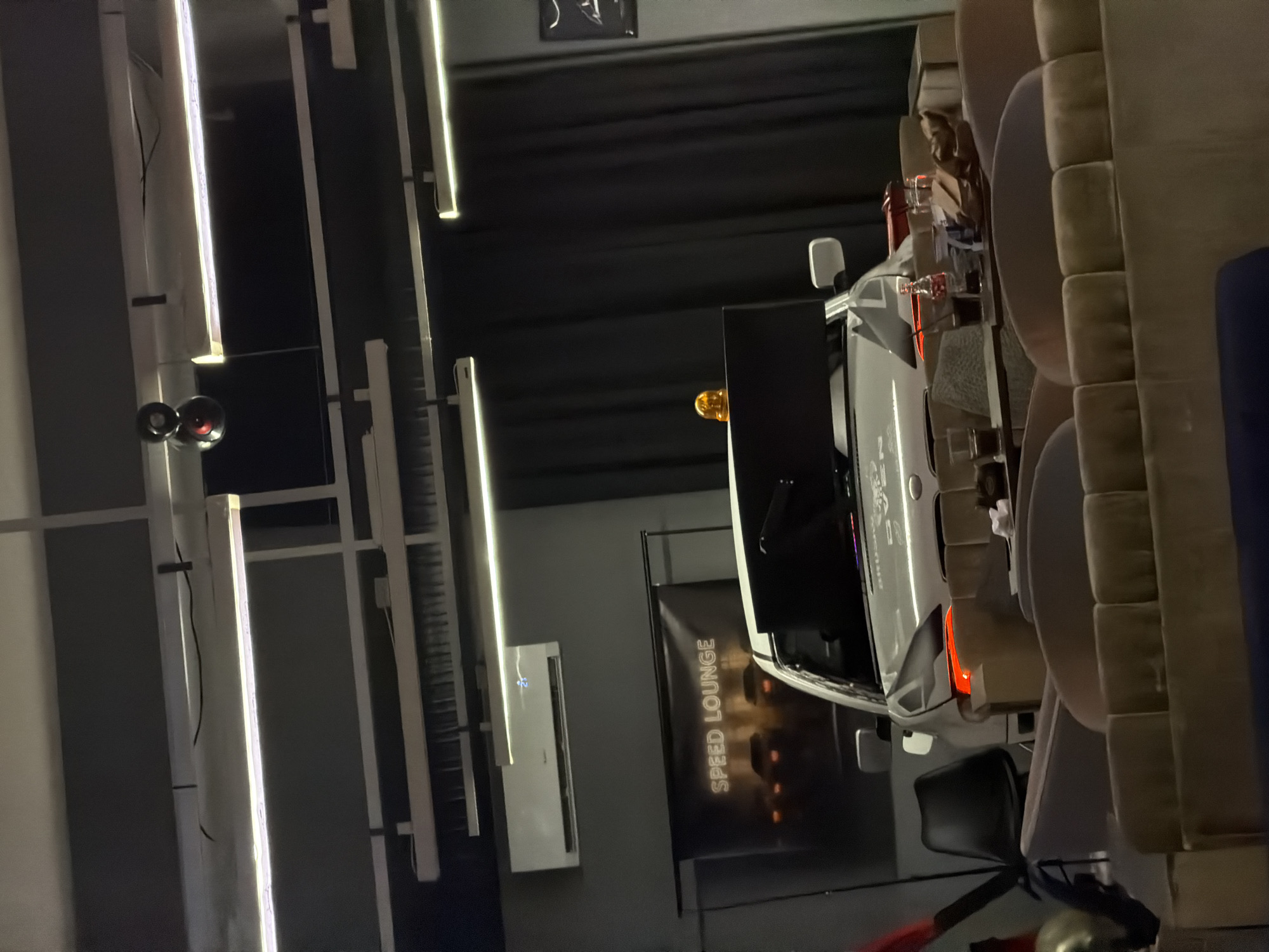 The image shows a dimly lit indoor space, possibly a lounge or a cafe, with a car as a central display. A banner with "SPEED LOUNGE" written on it is visible behind the car. The car is white, with its front end visible, and it has an orange light on top. There are several seating arrangements, including sofas and chairs, in the foreground. Drinks and some paper items are placed on tables in front of the seating. The environment appears modern with strip lighting fixtures on the ceiling. The image does not provide any specific indicators to pinpoint the location in Plovdiv, Bulgaria.