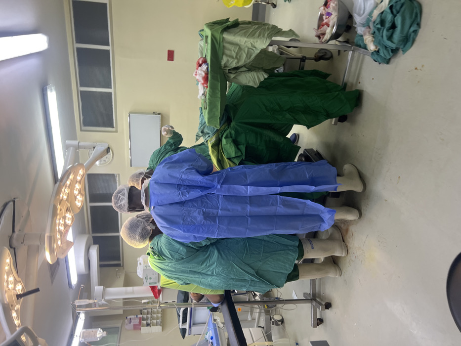 An operating room in Nairobi, Kenya, is depicted with at least four individuals present. Three individuals, wearing green and blue surgical gowns, scrub caps, and white protective boots, are positioned around a central surgical table covered with green drapes, indicating an ongoing medical procedure. One individual in a blue gown stands centrally with their back to the viewer, while another in a green gown leans over the table to the left. A third individual in green is partially visible to the right, also engaged with the table. Bright LED surgical lights are affixed to the ceiling directly above the surgical field.
To the left, an IV stand with a bag is visible. To the right of the central activity, a metal trolley holds surgical instruments and what appears to be tissue or surgical waste displaying reddish coloration. A pile of green surgical drapes is on the floor to the right, alongside a metal basin containing additional discarded surgical materials. The room features light-colored walls, high rectangular windows with horizontal blinds, and a light-colored floor. A wall clock and a small whiteboard are visible on the back wall. Some reddish-brown stains are present on the floor.