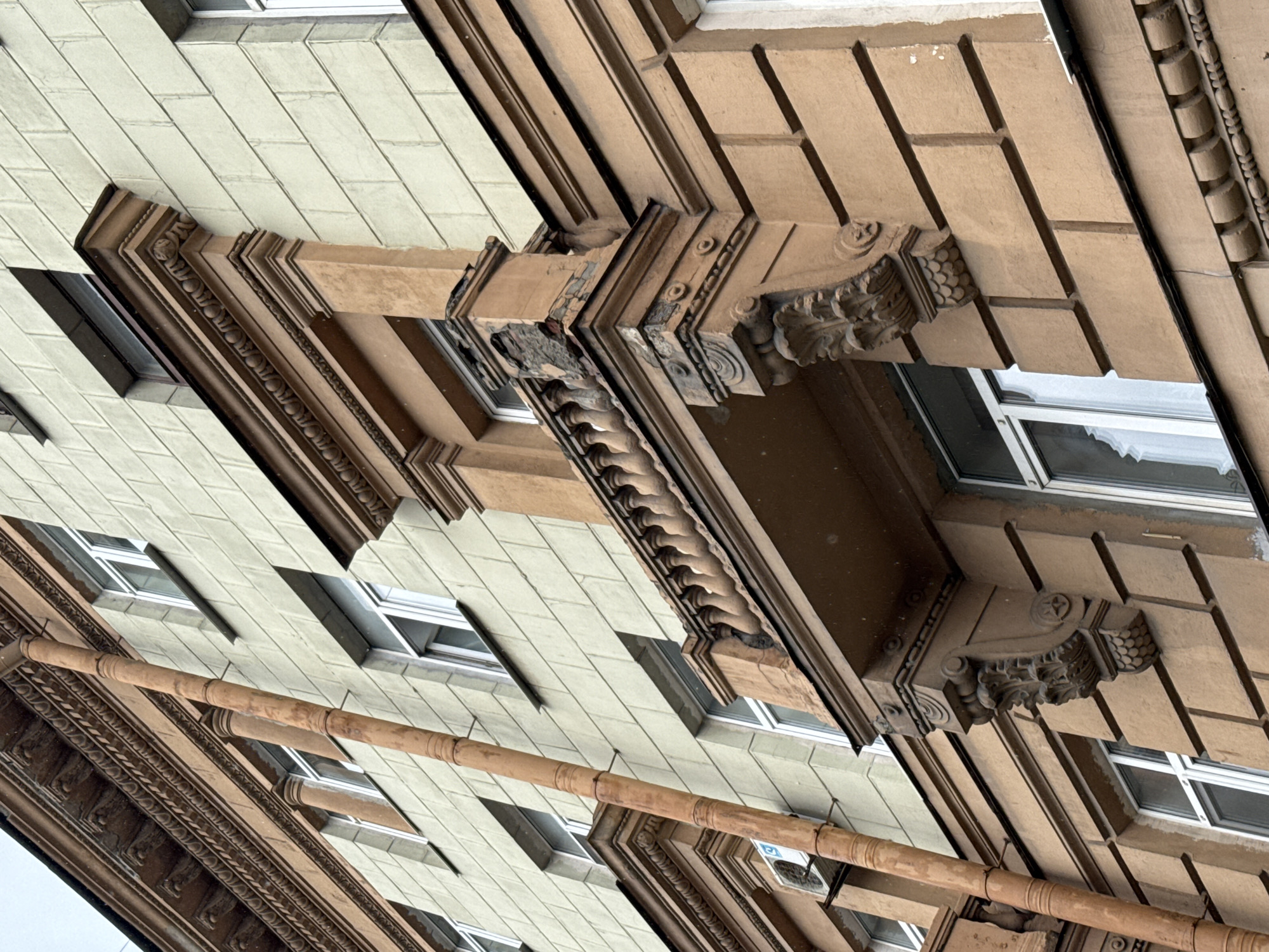 The image shows a close-up, low-angle view of a building facade in Saint Petersburg, Russia. The building appears to be old, with ornate architectural details. The facade is divided into two distinct color sections: a lighter, cream-colored upper section and a darker, reddish-brown lower section.
The upper section is characterized by rows of rectangular blocks, likely stucco or concrete imitating stone. It features several windows with white frames, some with visible white curtains inside. A long, weathered, reddish-brown pipe runs vertically down the facade, attached by several brackets. An air conditioning unit is mounted on the wall beneath one of the windows.
The lower section exhibits more elaborate decoration. A protruding balcony with a balustrade of small, round pillars is visible. The balcony is supported by large, carved corbels featuring acanthus leaf motifs and other decorative elements. The plaster on the balcony's edge and supports shows signs of damage and crumbling. This section also has more windows with white frames.
Above the cream-colored section, there is a band of decorative molding with intricate carvings, possibly a cornice. The overall impression is of a historic building that is showing signs of age and wear, with elements of classical or art nouveau architecture. The weather appears overcast, and there are small white specks in the air, suggesting either snow or light rain. No people are visible in the image.