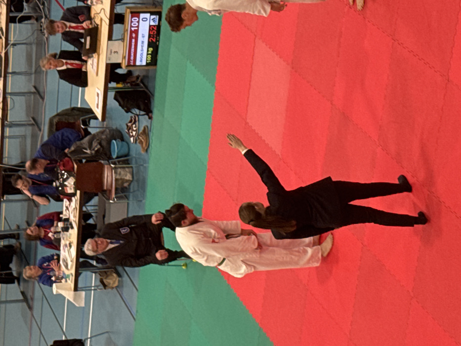 An indoor sports facility in Brumath, France, displays a combat sports event, likely judo. The scene features a competition mat composed of interlocking red and green sections. In the foreground, a female referee, dressed in a dark suit, stands on the red mat with her back to the camera, her right arm extended upward and outward, signaling. To her left, a male athlete in a white judogi with a green belt is bowing. To her right, another male athlete in a white judogi stands facing the first athlete. Both athletes are barefoot.
In the mid-ground, a male official in a dark suit is seated on a chair on the green mat. Behind him, multiple individuals are seated at two long tables. The table on the left hosts four individuals, two female and two male, with various items including bottles and papers. The table on the right features two officials, one female and one male, seated with a laptop open. Below this table, a digital scoreboard indicates "A. C. QUATZENHEIM - 67 100" and "LINGOLSHEIM - 67 0," with "100 kg" and "2:52" displayed, alongside an Adidas logo. Personal belongings, including shoes and bags, are visible on the green mat around the tables. The area is brightly lit.