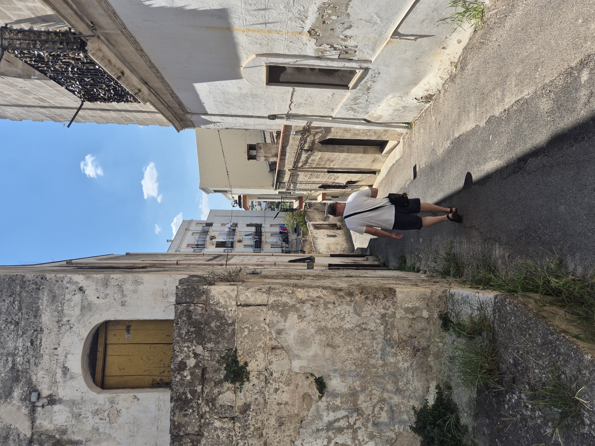 A person walks down a narrow, paved alleyway in Laterza, Italy. The person is wearing a white t-shirt, dark shorts, and sandals, and carries a black shoulder bag. The alley is bordered by old, weathered stone buildings on both sides. The building on the left has a rough, textured wall with patches of peeling paint and a yellow arched doorway with a wooden door. Green vegetation grows in cracks and along the base of the walls. The building on the right has a lighter-colored, textured facade with a dark rectangular window. In the background, other buildings and balconies are visible, with a bright blue sky overhead dotted with a few white clouds. The scene suggests a sunny day in a historic urban environment.