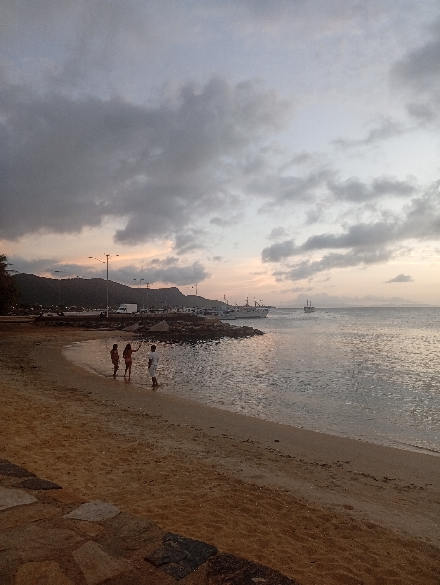 The image depicts a serene beach scene at dusk, with a cloudy sky casting a muted light over the landscape. In the foreground, three individuals are standing in shallow water near the shore. Two are facing away from the camera, and one is partially turned to face it, with an arm raised as if waving or posing. The sand on the beach is a warm, light brown, with a pathway of irregular stones leading into the foreground. The water is calm and reflects the soft, diffused light. In the middle ground, a rocky embankment separates the beach from a harbor area. Several boats are docked or anchored in the water, with a larger white boat and a smaller, older-looking vessel visible. Beyond the harbor, a mountainous silhouette is visible against the horizon, partially obscured by the twilight. Streetlights are scattered along the shoreline, and a white truck is parked near the harbor. The overall atmosphere is peaceful and tranquil.