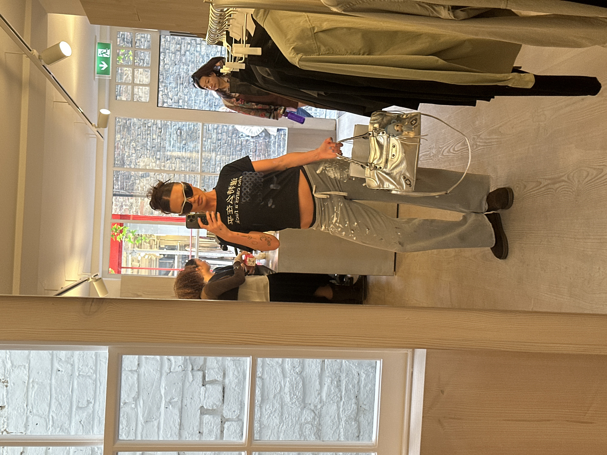 A reflective image in a shop, presumably in London, United Kingdom, shows a person posing with a silver handbag while holding a phone as if taking a picture. Another person stands in the background near hanging clothes. The environment features brick-walled buildings visible through windows, suggesting an urban setting. The light fixtures suggest a modern interior.