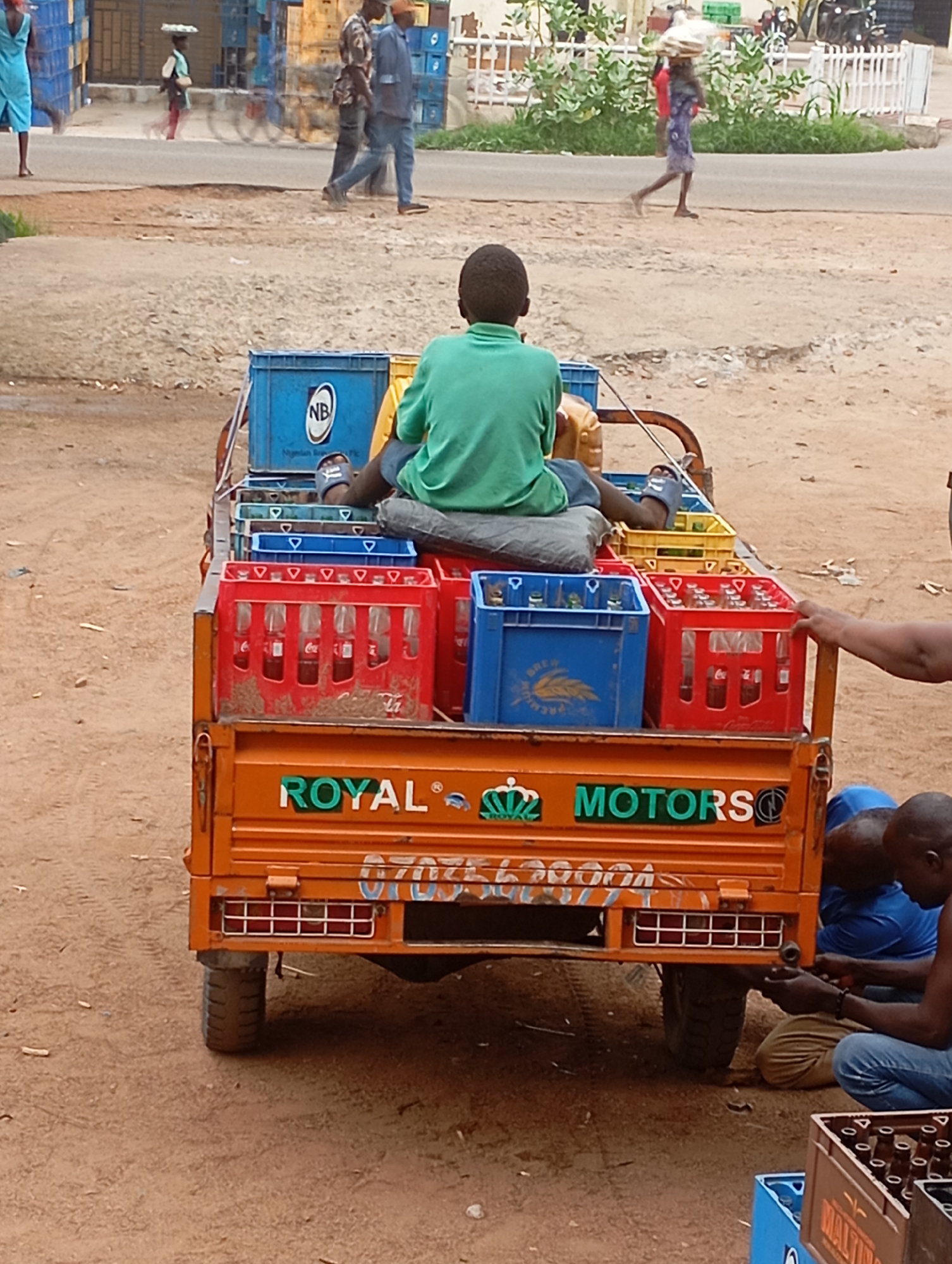In Shendam, Nigeria, a young boy in a green shirt sits atop a bright orange three-wheeled vehicle laden with crates of beverages. The vehicle, emblazoned with "ROYAL MOTORS," is parked on a dusty, reddish-brown road. Several crates, some red and some blue, are stacked on the back of the vehicle, filled with glass bottles, many appearing to be Coca-Cola. Two men, one in a blue shirt and the other in a black shirt, are crouched behind the vehicle, appearing to be working on it or its cargo. In the background, other individuals are visible, some walking and others standing near buildings. A woman in a blue dress walks in the distance, and other people are seen near a white fence. The overall scene depicts a typical street-level commercial activity within a Nigerian city.