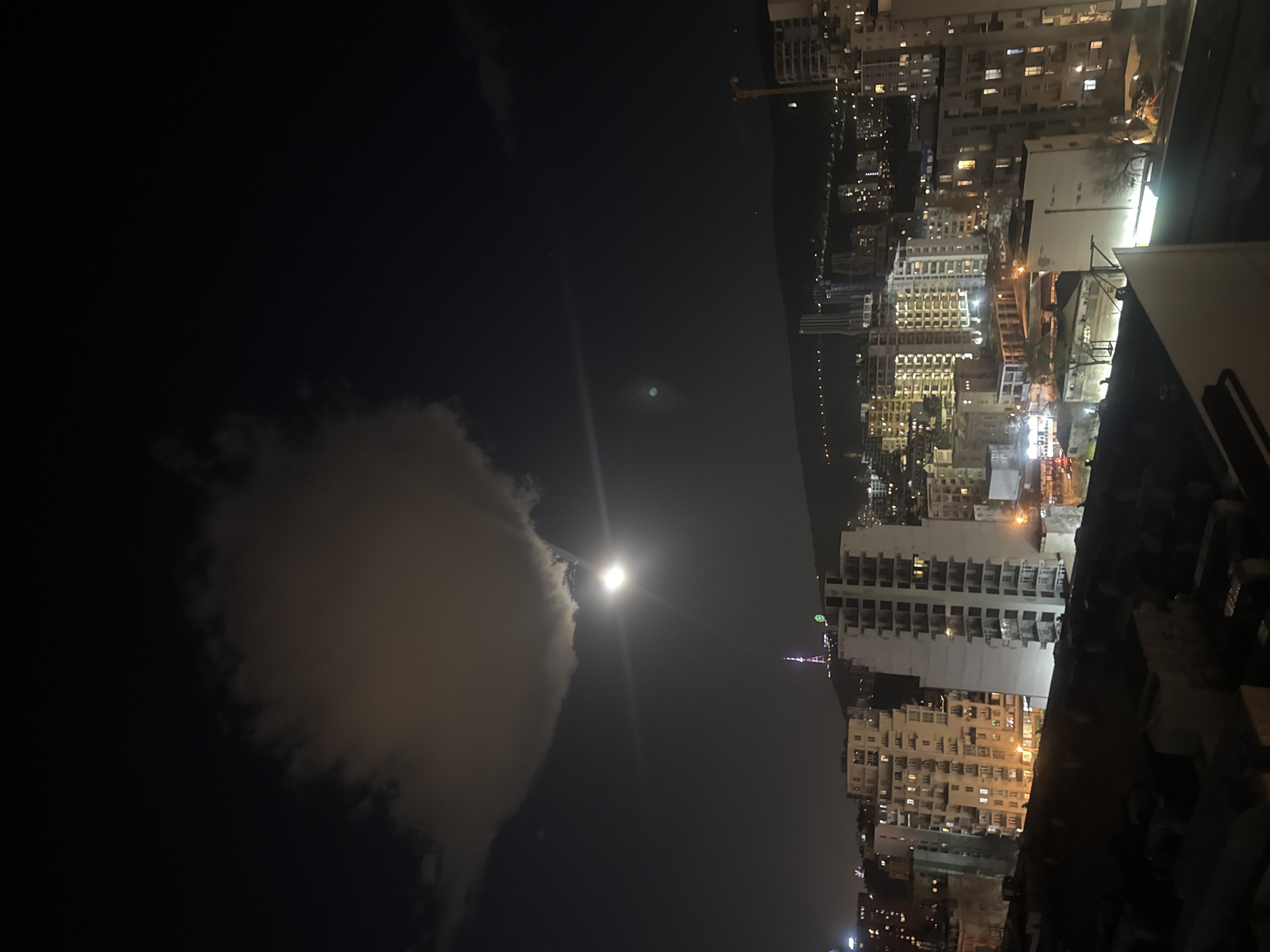 The image depicts a nighttime cityscape, likely taken from a high vantage point such as a rooftop. The skyline is dominated by illuminated buildings, suggesting a densely populated urban area. A body of water is visible in the distance, perhaps a river or bay. The sky is dark, with a bright moon obscured by clouds. A distant tower-like structure is visible among the buildings, potentially resembling a telecom or broadcast tower with colorful decorative lighting. This scene is likely located in Tbilisi, Georgia, owing to the unique appearance of the structure that may be the Tbilisi TV Broadcasting Tower, which is often illuminated at night. There are no people visible in this image. The environment is urban and nocturnal, and the image is taken at nighttime.