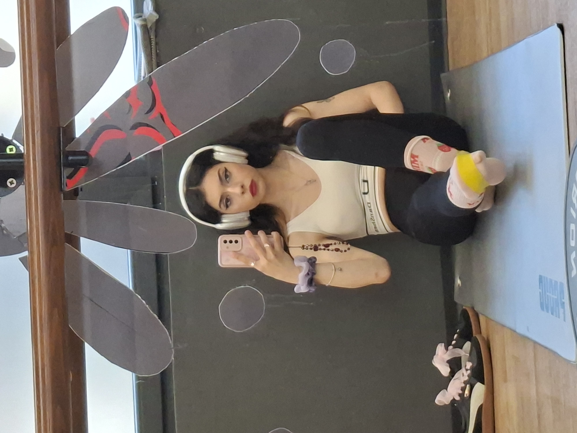 A young woman is captured in a mirror selfie, lying on her back with her head towards the left of the frame. She has long, dark, wavy hair and is wearing white over-ear headphones. Her face features prominent eyeliner and red lipstick. She is dressed in a white crop top with "DEUZIQ" visible on the elastic band and black leggings. On her feet, she wears light pink socks with strawberry patterns and the partial text "MOM" visible, one sock also featuring a bright yellow band.
She holds a light pink smartphone in her left hand, taking the photograph. Her right arm rests across her body, adorned with a beaded bracelet and a purple scrunchie on her wrist. Rings are visible on her fingers.
The setting appears to be indoors, possibly a gym or a room designed for exercise, located in Sofia, Bulgaria. The background, as reflected in the mirror, is a dark, possibly grey, surface decorated with various abstract light grey oval and circular shapes. On the far left, a wooden vertical beam or frame is seen, next to which are decorative grey cutouts with red and black abstract patterns, resembling dragonflies or similar motifs.
To the right of the woman, a light blue/grey yoga mat is laid out on a light wooden floor. The brand name "PUMA" is partially visible and rotated on the mat. Next to the mat, a pair of black sneakers with white soles and light pink laces are also resting on the wooden floor. The lighting suggests an indoor environment, likely during the day or in a well-lit space. The woman's expression is neutral as she poses for the picture.