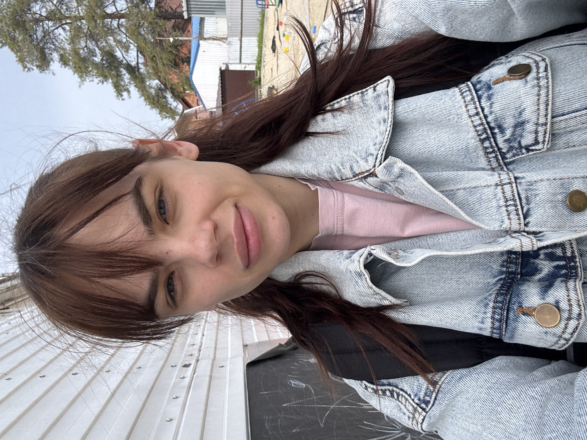 A young woman is featured in a selfie, looking directly at the camera with a subtle, closed-lip smile. She has long, dark brown hair with bangs and light-colored eyes. She is dressed in a light-wash, acid-washed denim jacket with bronze buttons, worn over a light pink top. A black strap, likely from a bag or backpack, is visible beneath her jacket.
The photograph was taken outdoors in Rostov-on-Don, Russia, during daytime under natural light. The background is softly focused, showing an urban or suburban environment. Above her, a large tree with green foliage reaches towards a light, possibly overcast, sky. Behind her, buildings, including some with blue roofs, and wires or clotheslines are discernible. To her right, a white corrugated metal wall or fence provides a textured backdrop. Below her, a dark, flat surface with white, chalk-like markings or scratches is visible. The woman's expression conveys a calm and pleasant demeanor. There is no visible text in the image.