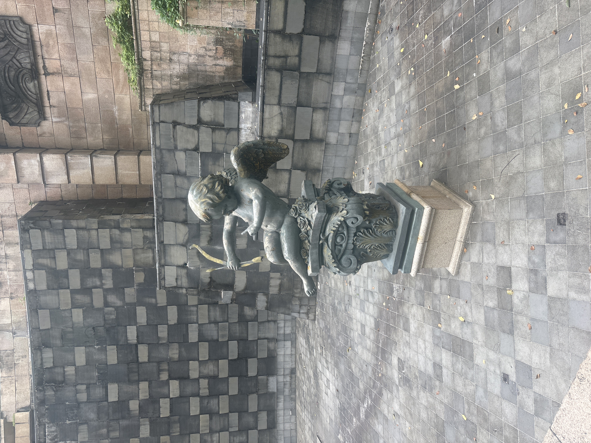 An outdoor shot captures a bronze statue of a cherub holding a bow and arrow, positioned on a pedestal. The cherub is seated, with its left leg bent and right leg extended. Its wings are spread, and its head is turned to the left. The statue sits atop a decorative, ornate base featuring floral motifs. The statue and its base are set against a backdrop of a large, stepped wall constructed of stone blocks. The ground in front of the statue is paved with square tiles arranged in a diamond pattern. Scattered leaves are visible on the tiled ground. The overall atmosphere is serene and somewhat aged, suggesting it is located in a public space or garden within the Nanhai District of China.