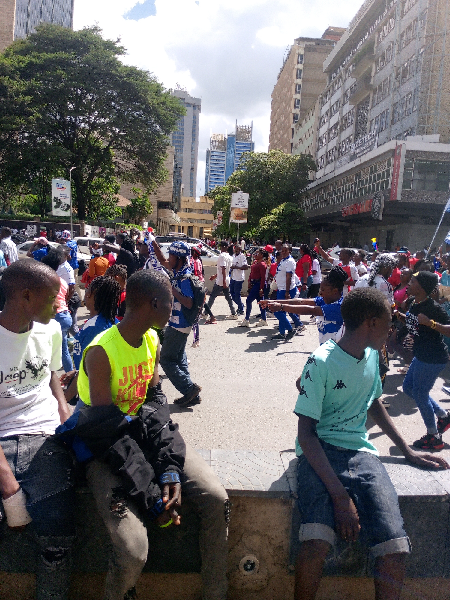 The image shows a street scene in Nairobi, Kenya, with a crowd of people gathered for what appears to be a march or demonstration. There are several tall buildings in the background, including what appears to be the Zetech University building and modern skyscrapers.
In the foreground, three young men are sitting on a low wall or ledge. The one on the left is wearing a white Jeep-branded T-shirt and dark jeans. The middle one is wearing a yellow tank top with a "Just Kill" print and ripped light-colored jeans with a black jacket draped on his lap. The third young man is wearing a light blue Kappa T-shirt and dark denim shorts.
Behind the young men, a diverse crowd is filling the street. Most are dressed in casual clothing, with some wearing red, white, or blue garments, possibly indicating support for a particular cause or team. Some individuals are carrying flags or waving their arms, suggesting active participation in the event.
Advertisements and signs are visible in the background, including one for an Aristo Beef Burger, RIC brand tires, and AFC Leopards. The presence of these elements further contextualizes the scene as taking place in a busy, urban area.