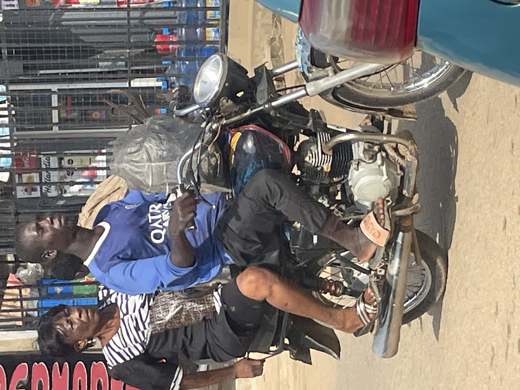 A medium shot captures two individuals, a woman and a man, riding a motorcycle on a dusty road. The woman is seated behind the man. The man is wearing a blue t-shirt with the "QATAR AIRWAYS" logo and black pants. The woman is wearing a black and white striped top and black shorts, and carrying a bag with the "FENDI" logo. The motorcycle is dark colored with a silver exhaust pipe. In the background, a store with a metal grate displays bottled water and various other products. The scene appears to be on a street in Abuja, Nigeria, given the context of the image's origin.