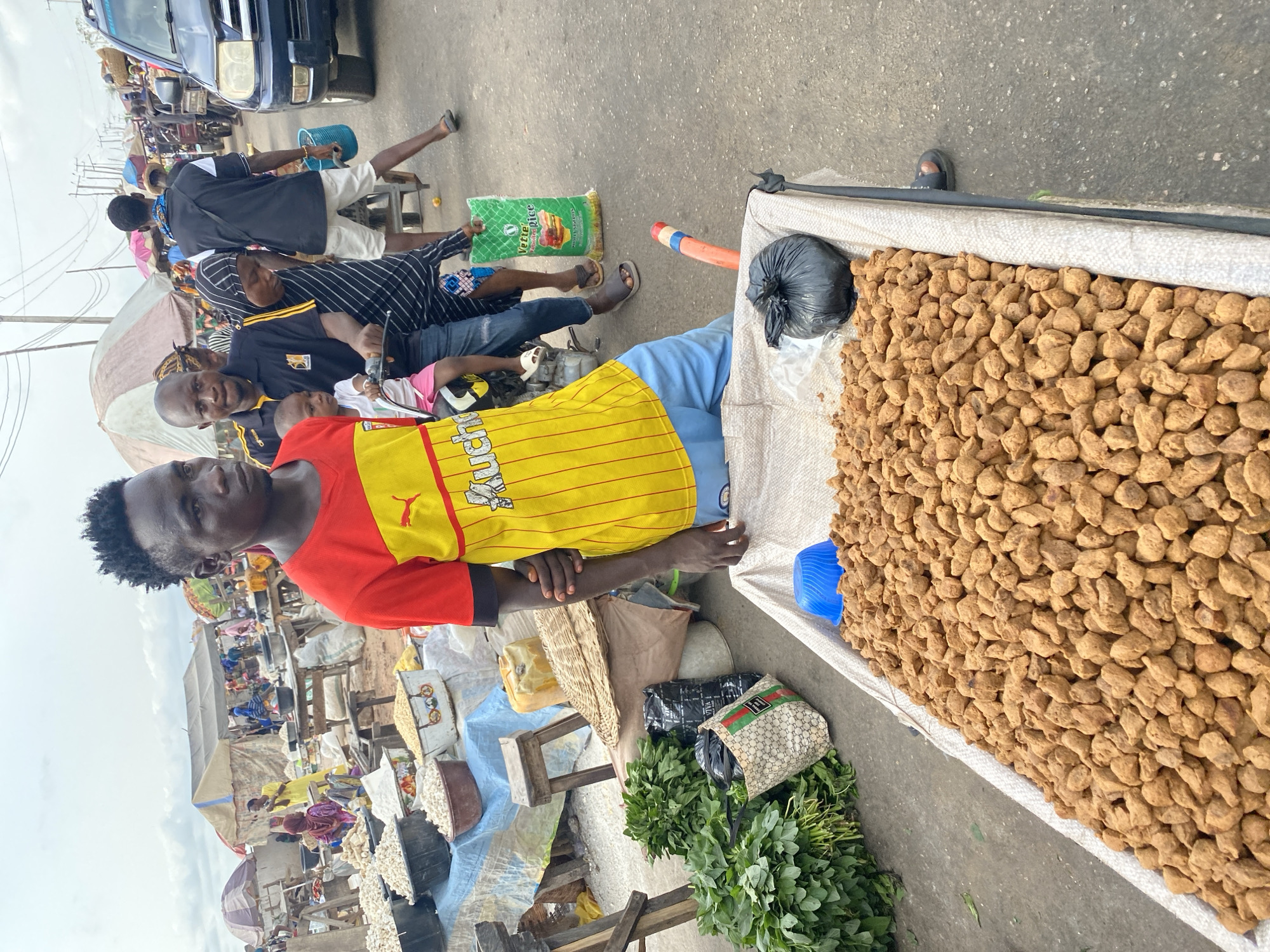 The scene depicts a bustling marketplace in Lokoja, Nigeria. A man in a red and yellow soccer jersey stands by a cart filled with what appear to be small, brown, pebble-sized food items, possibly groundnuts or a similar type of snack. Beside the cart, there are bags of leafy green vegetables and other containers, suggesting a variety of produce for sale. Other individuals are present in the background, including a man holding a child, people walking, and cars parked nearby. Market stalls and umbrellas create a densely packed environment under an overcast sky.