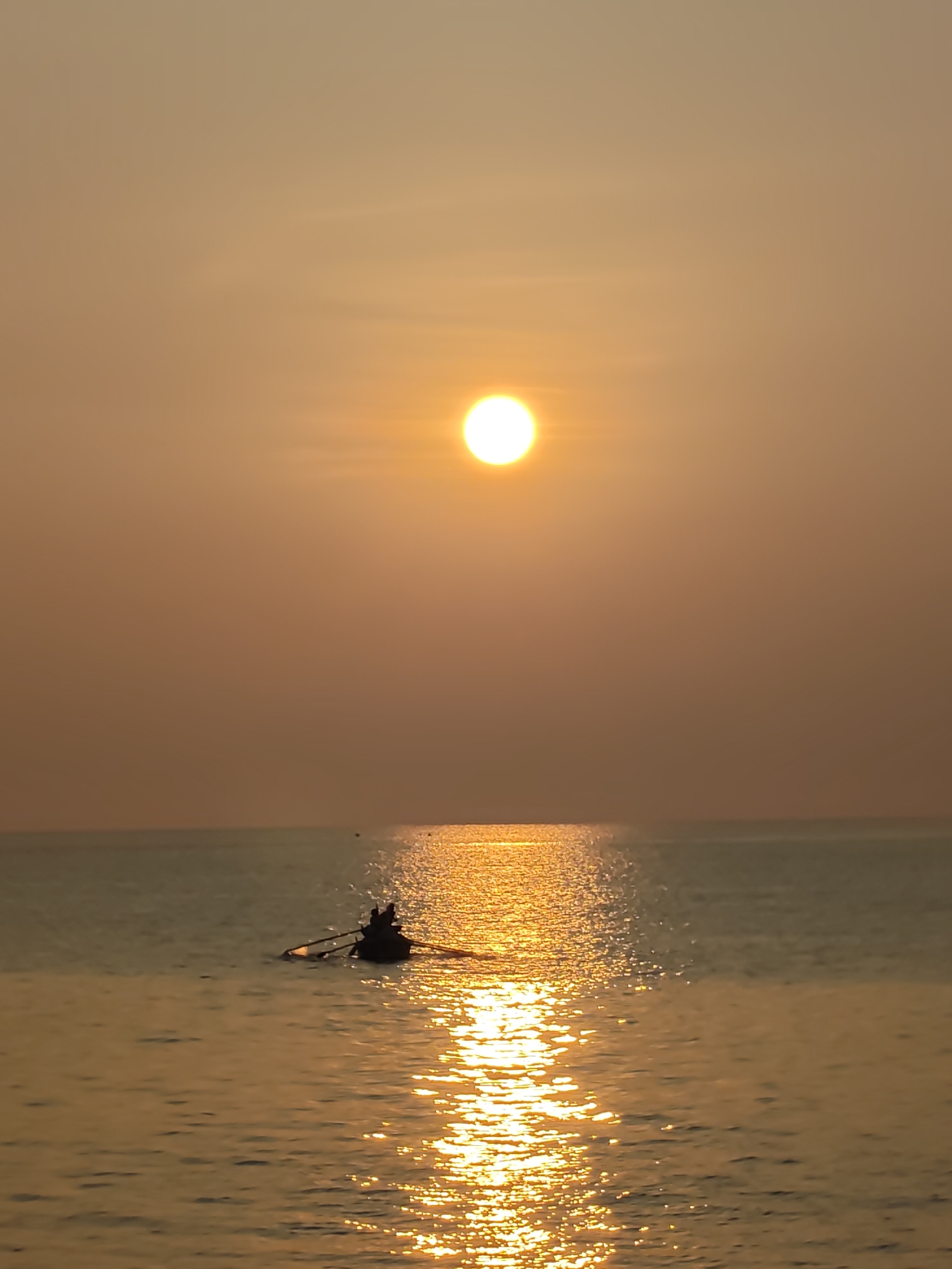 The image shows a silhouetted rowboat with two occupants on a calm body of water, likely the sea, under an orange sky dominated by a bright, low sun. The water reflects the sun's light, creating a golden path stretching towards the viewer. The scene appears to be taking place at sunrise or sunset. The location is presumably Lhokseumawe, Indonesia, given the prompt's details, but the image itself doesn't offer definitive geographical clues.