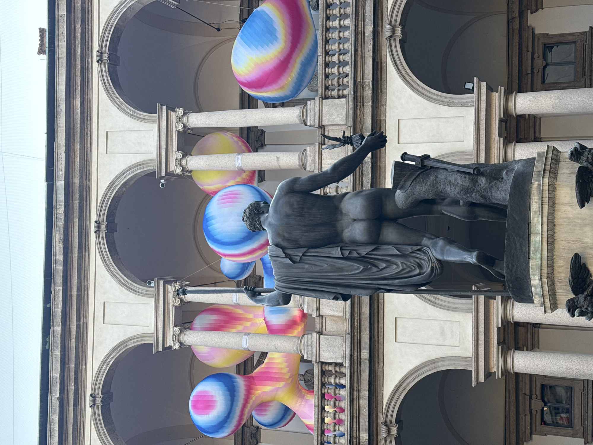 An image, originally captured in portrait orientation but rotated on its side, displays a classical statue and colorful art installations within a grand architectural setting.
In the foreground, a dark, weathered statue of a male figure stands prominently. He is depicted from the back, nude to the waist, with a draped garment covering his lower body. His right arm is raised, holding what appears to be a staff or sword. The statue rests on a pedestal, which features a dark, bird-like sculpture on one side.
Behind and around the statue, a series of large, inflatable, balloon-like objects are suspended from the ceiling of an arcade. These installations are characterized by vibrant, swirling patterns of blue, pink, yellow, and white stripes. They vary in shape, with some being spherical and others more elongated, creating an abstract and playful contrast with the classical architecture.
The background consists of a stately stone building featuring a repeated pattern of arches supported by columns, indicative of a colonnade or arcade. The stone is light in color, possibly white or cream, and shows signs of age. Dark windows are visible on the upper levels of the building. The overall lighting suggests it is daytime, likely overcast, as there are no harsh shadows. There is no visible text in the image. The scene likely depicts an outdoor art installation or exhibition in a historical location, consistent with the provided context of Milan, Italy.