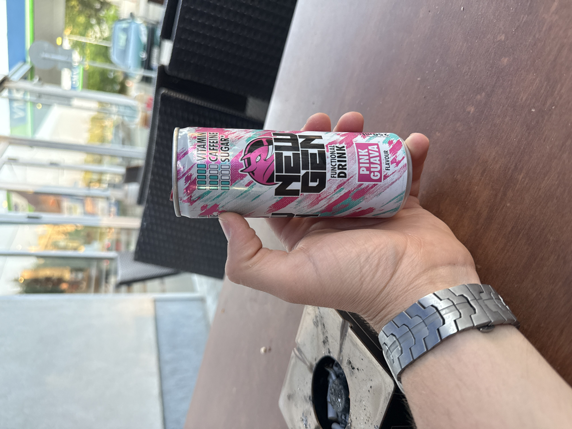 A first-person perspective photograph depicts a right hand holding a silver and white "NEW GEN" brand functional drink can, featuring a pink devil head logo and "Pink Guava" flavor. The can's design includes diagonal pink and teal brush strokes and prominently indicates the presence of "VITAMIN" and "CAFFEINE" (represented by five filled green squares each) and the absence of "SUGAR" (represented by five empty white squares). The hand wears a silver-toned metal watch with a segmented, angular band on the left wrist. The scene is set on a dark brown wooden table, which exhibits wear and minor debris. To the left, a square, dark grey metallic ashtray contains ash and several discarded cigarette butts.
In the midground, blurred dark-colored outdoor chairs are visible. The background, viewed through large glass doors and windows, shows an outdoor area. Reflections in the glass suggest an interior space. Beyond the glass, a brightly lit gas station is discernible, identified by a green and blue striped canopy with partial lettering ("VIK") and a blurred dark green car in a parking area. The overall lighting suggests a clear day. The location for this scene is Stara Zagora, Bulgaria.
