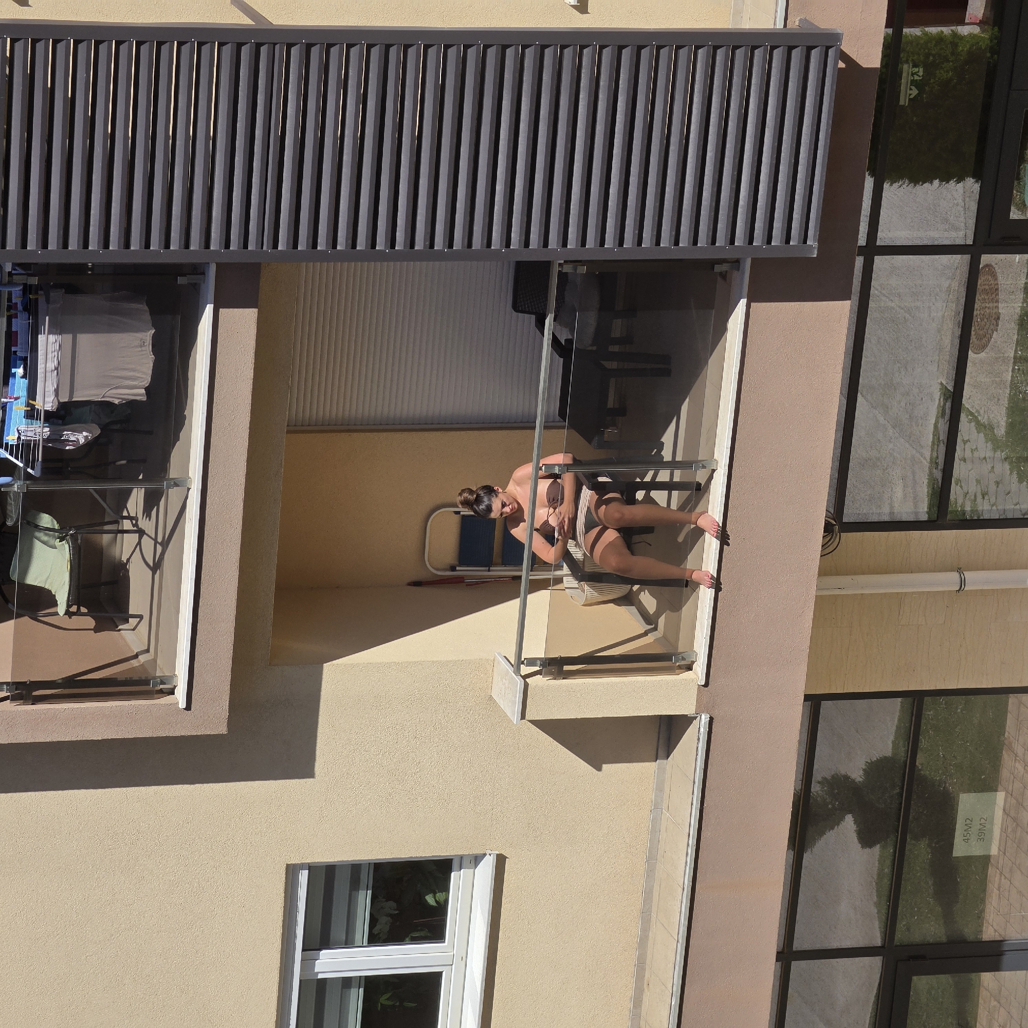 A rotated medium shot depicts a building with multiple balconies in Podgorica, Montenegro. On one of the balconies, a woman in a bikini is sitting on a chair, presumably tanning. Other balconies show signs of habitation, such as clothes drying on a rack and furniture. The building's exterior is painted in light and dark shades of beige. A window is visible on the lower left of the image, and a large window reflecting the exterior view of greenery and a walkway is seen on the right.