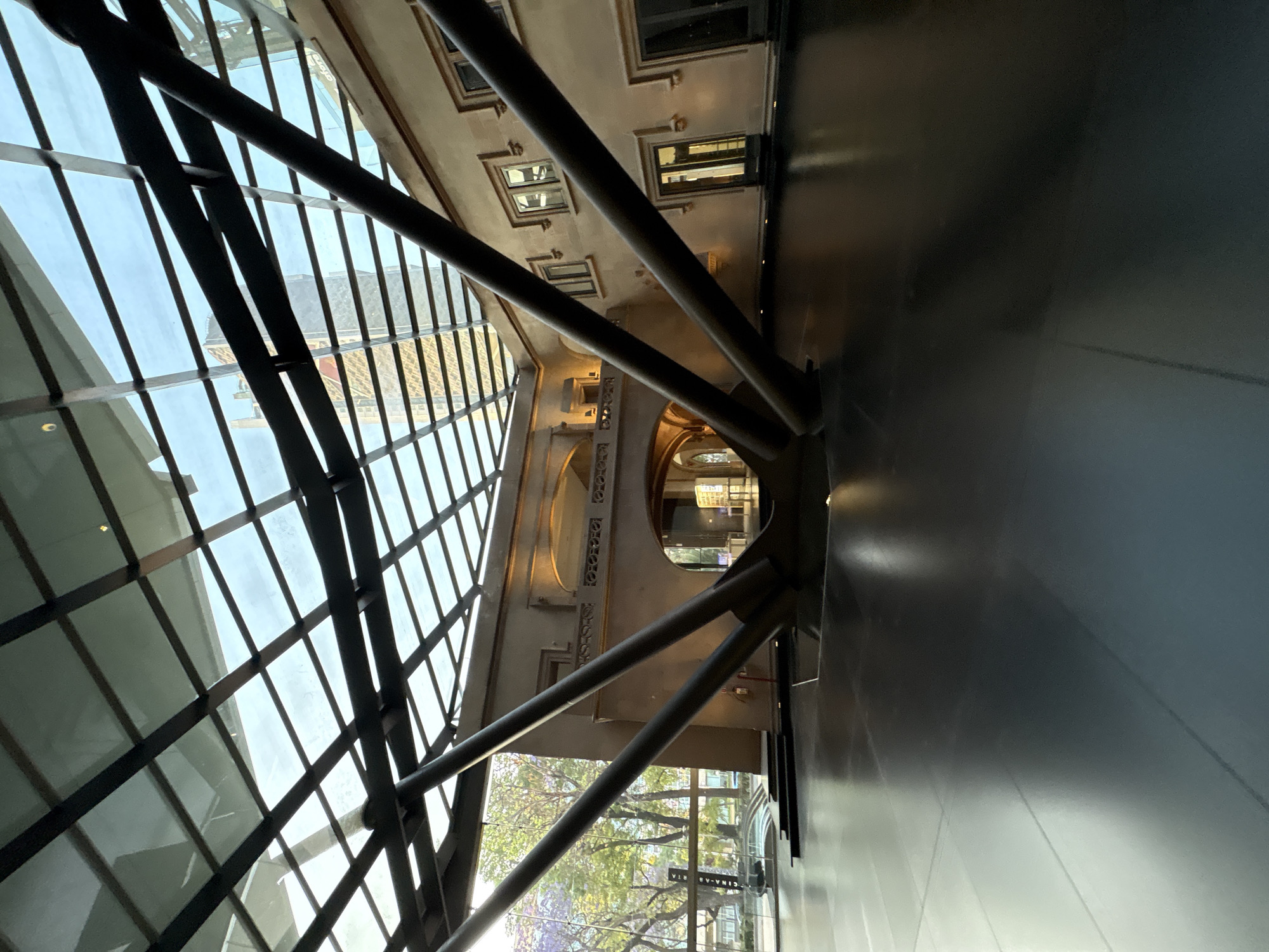 This image depicts the interior of the Centro Cultural Elena Garro in Coyoacán, Mexico City, Mexico. The scene is an expansive architectural space featuring a large, multi-paneled glass roof supported by a grid of dark, rectangular metal beams. A prominent structural element consists of several thick, dark, cylindrical steel beams that converge from the roof towards a central point on the reflective floor, splaying outwards diagonally.
Beneath this modern glass and steel canopy, a two-story, light-colored historical building facade is enclosed. This facade features multiple rectangular windows with dark frames on both levels, a decorative balustrade balcony on the upper level, and two arched passages on the ground floor. Artificial lighting illuminates sections of the facade, particularly above the arches and along the balcony.
The floor is a dark, smooth, and highly reflective surface, mirroring the overhead glass structure and interior lighting. Through the large glass panels on the left side, an exterior view reveals lush green trees interspersed with purple-blossomed trees, indicative of an urban park or street environment. A tall, modern building with a light-colored facade is partially visible and reflected through the upper right section of the glass roof. No individuals are present in the scene. A red fire extinguisher is mounted on the wall near the left arched passage. A sign, partially obscured, displaying "CINEMAX" and additional text, is visible through the left glass wall.