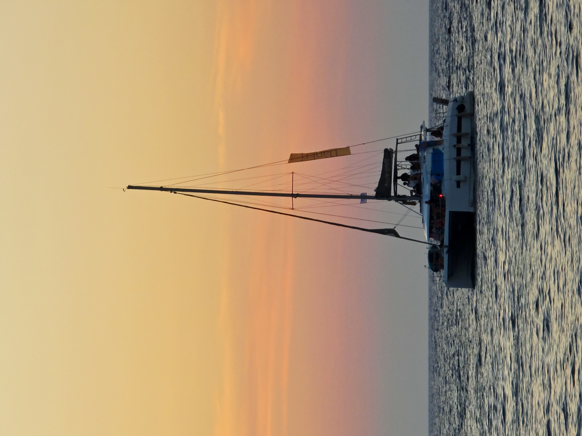 A white catamaran is sailing on the sea at sunset. The sky is a gradient of yellow, orange, and pink. The catamaran has multiple people on board and a tall mast with sails. There are waves on the water. The scene is calm and serene. The location is the city of Chania, Greece.