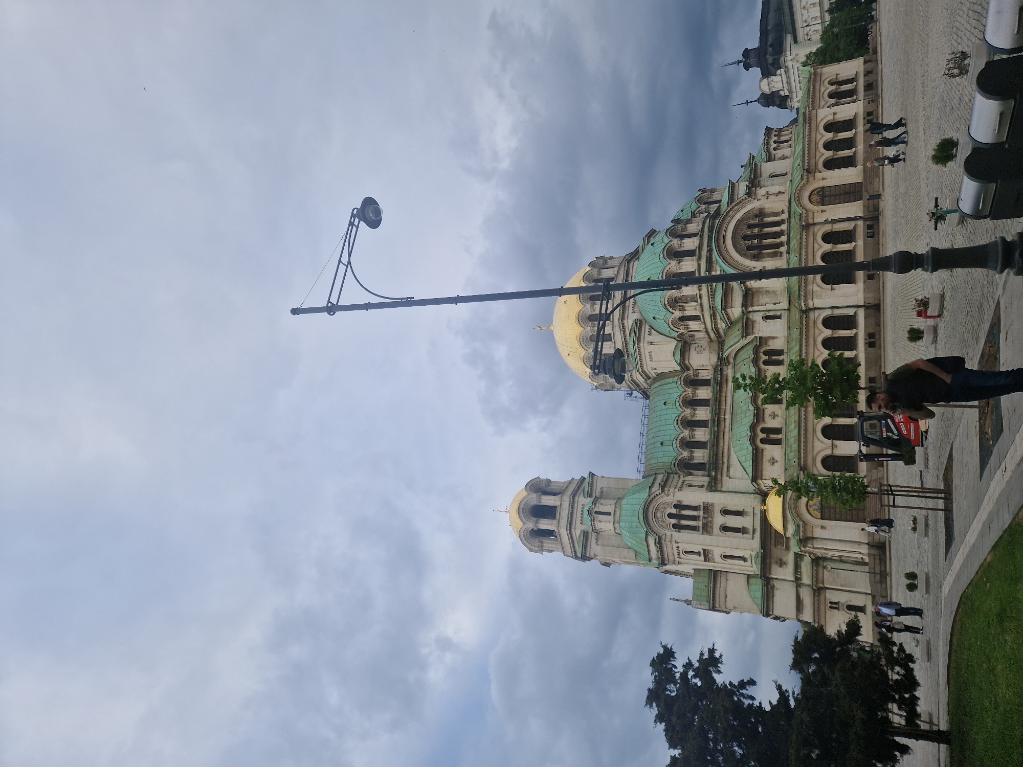 The image depicts the Alexander Nevsky Cathedral in Sofia, Bulgaria, framed by a cloudy sky and a street lamp. The cathedral's intricate architectural details, including its domes and facades, are visible. People can be seen walking on the pavement in front of the cathedral. A tree is positioned in the lower right corner of the frame.