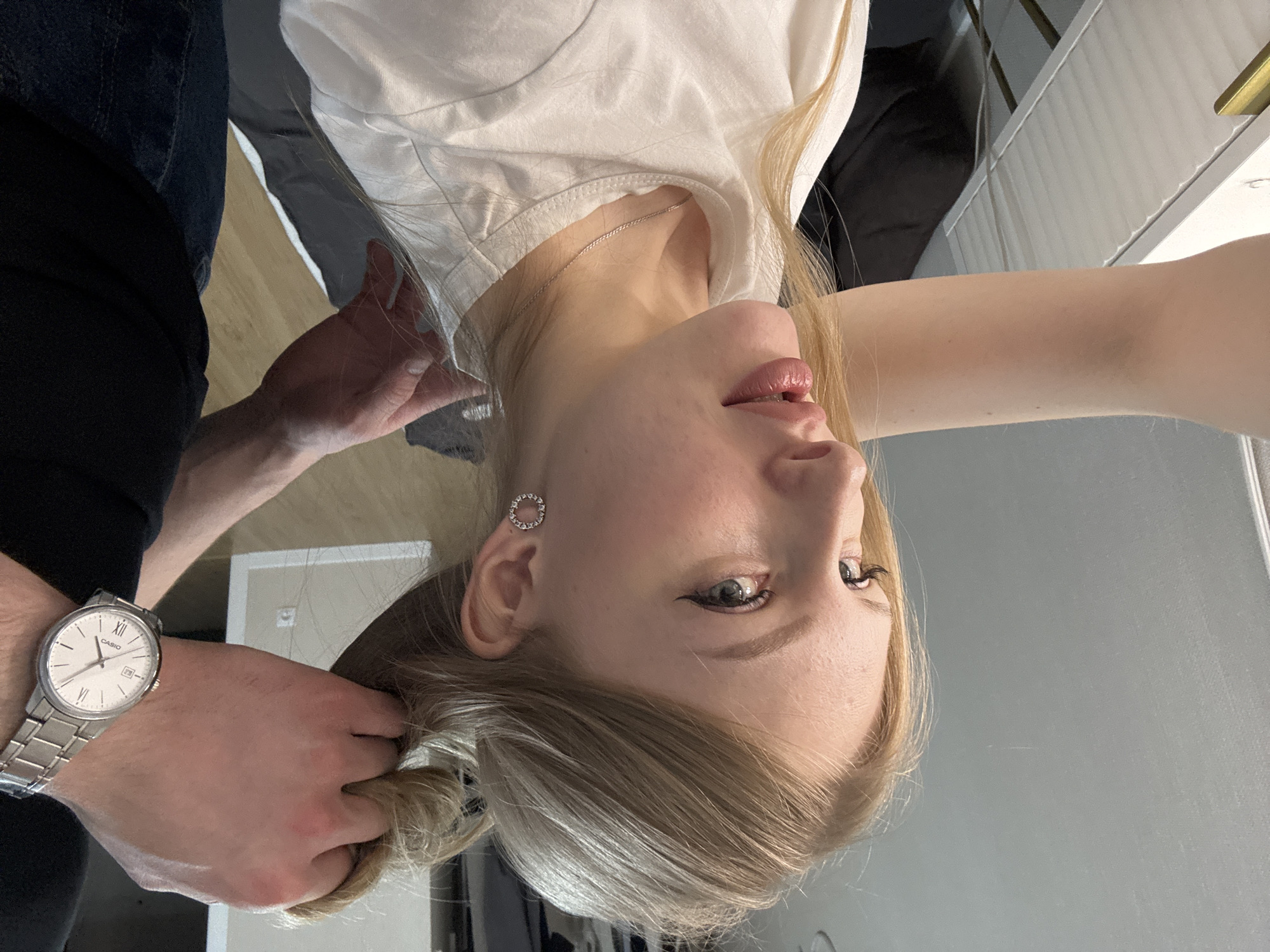 This portrait, captured in an indoor setting in Perm, Russia, features a young blonde woman having her hair styled. Her head is tilted back and positioned centrally, looking slightly upwards and to the right with a calm expression. She has fair skin, light-colored eyes accented with winged eyeliner and mascara, and her lips appear glossy. She wears a simple white t-shirt and a delicate silver chain necklace. A notable detail is the circular, open-work silver earring in her left ear.
On the left side of the frame, the arm of another person is visible, clad in a black top or sleeve and dark blue jeans. This person's hands are gently gathering or working with the woman's blonde hair. A silver-toned Casio watch, featuring a white face with Roman numerals "VI" and "XII" prominently displayed, adorns their left wrist.
The background shows a mix of light and dark surfaces, suggesting interior walls or furniture. A light wooden floor is visible below the woman's head (when oriented correctly), and some white paneling with a gold accent is subtly present. The lighting is bright and even, indicating a well-lit indoor environment, possibly a home or a salon, where a personal grooming or hairstyling session is taking place.