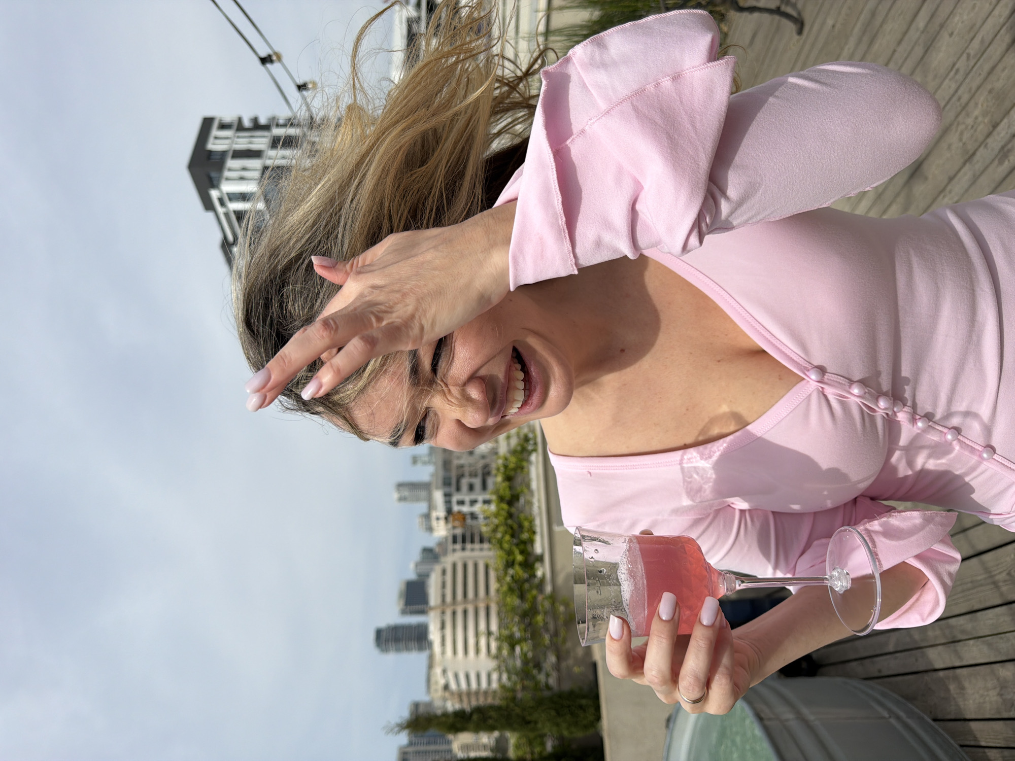 A smiling woman with blonde hair, wearing a pink top, holds a glass of pink liquid in front of her. She has one hand covering her face. The background shows a city skyline under a pale sky, with buildings of varying heights and designs. The woman is standing on a wooden surface, likely a deck. The location is Tel-Aviv, Israel.
