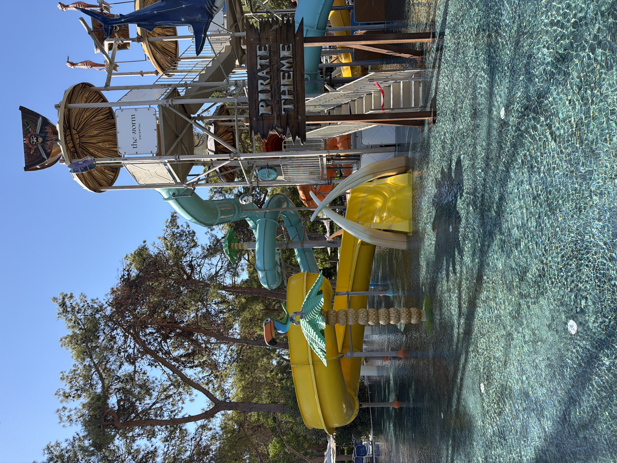 The image depicts a section of a water park in Kemer, Turkey, specifically a "Pirate Theme" area. The foreground shows a shallow pool with clear, rippling turquoise water. In the mid-ground, a large yellow water slide curves into the pool, and next to it are decorative elements resembling mammoth tusks. Further back, a multi-level water play structure is visible, featuring various colorful slides (teal and orange), a shark figure, and nautical-themed decorations including seahorses and a skull banner. A sign with "PIRATE THEME" in distressed wood lettering is prominently displayed. The background is dominated by tall pine trees against a clear blue sky. No people are visible in the scene. The overall impression is of a vibrant and engaging children's play area within a resort setting.