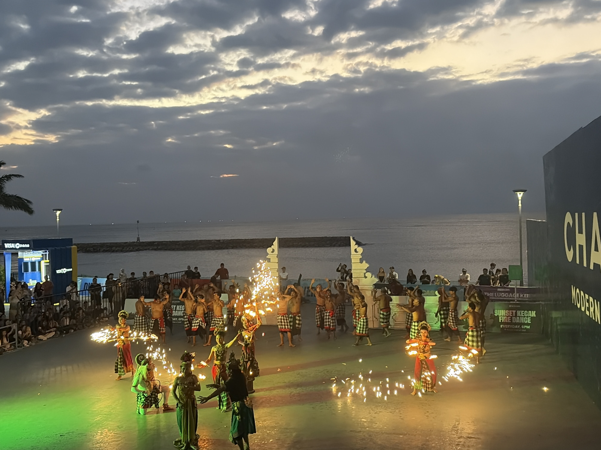A group of Balinese dancers perform a Kecak fire dance at sunset on a paved outdoor stage in Kuta, Indonesia. The dancers are wearing traditional attire, including sarongs and headdresses. Some of the dancers are holding torches or props that are emitting sparks, creating a dramatic visual effect. The performance is taking place on a stage with green lighting on the left side. In the background, there is a large body of water, likely the ocean, and a cloudy sky with the sun setting. A crowd of spectators is visible watching the performance. There are also some structures and signage in the background, including what appears to be a VISA ATM and a banner that reads "SUNSET KECAK FIRE DANCE". The overall atmosphere is one of a lively cultural event.