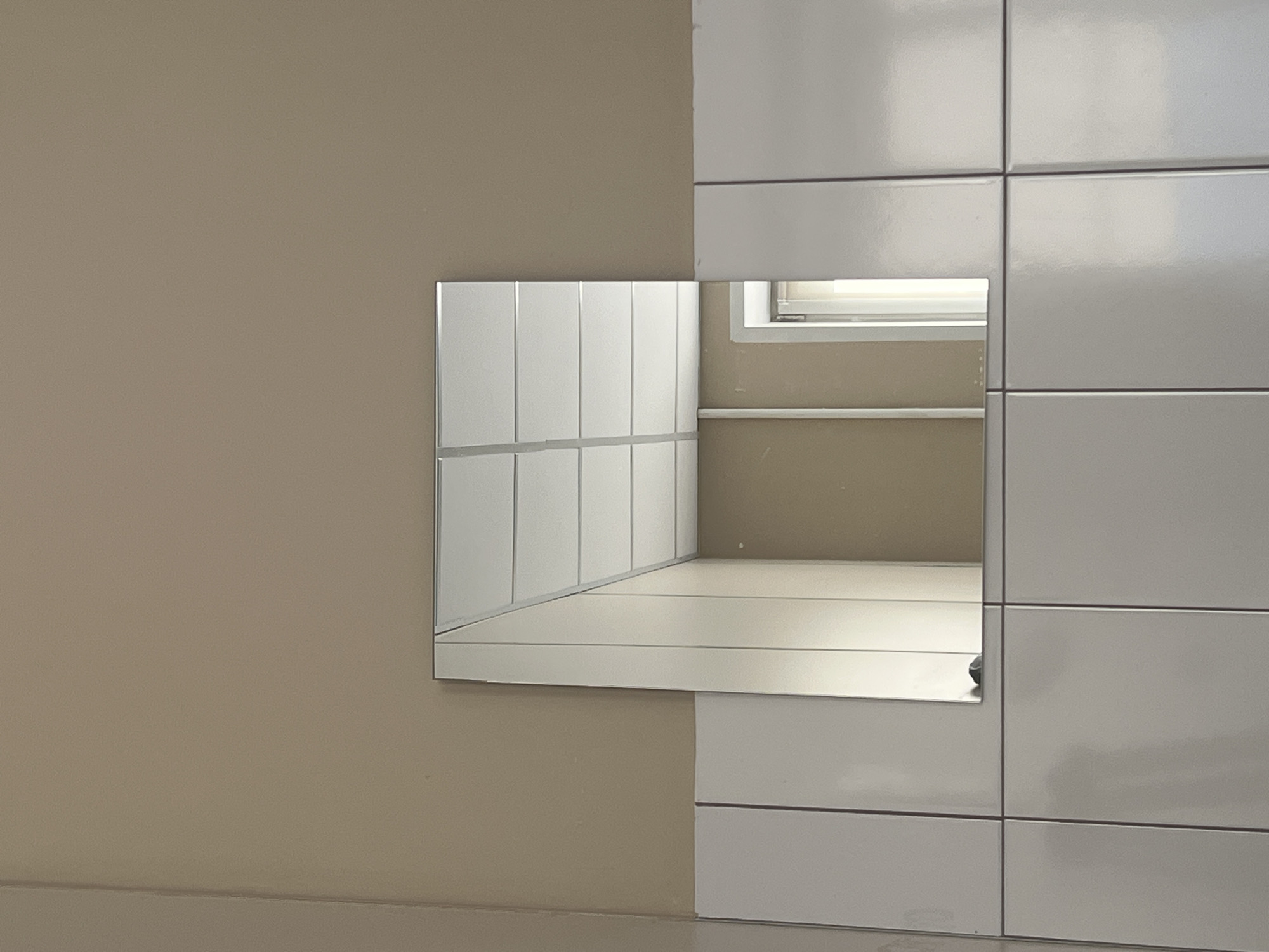 The image depicts a square mirror mounted on a beige wall. Below the mirror, a tiled surface is visible, consisting of rectangular white tiles with dark grout lines. The mirror reflects a section of a dropped ceiling, characterized by its grid of white panels, and a hallway with beige walls. A doorway or opening is also partially visible in the reflection, suggesting an interior space. There is a small, dark object at the bottom edge of the mirror, possibly a stain or a small item. The image appears to be taken indoors, likely in a public or commercial setting such as a restroom or corridor. The location is specified as Khimki, Russia.