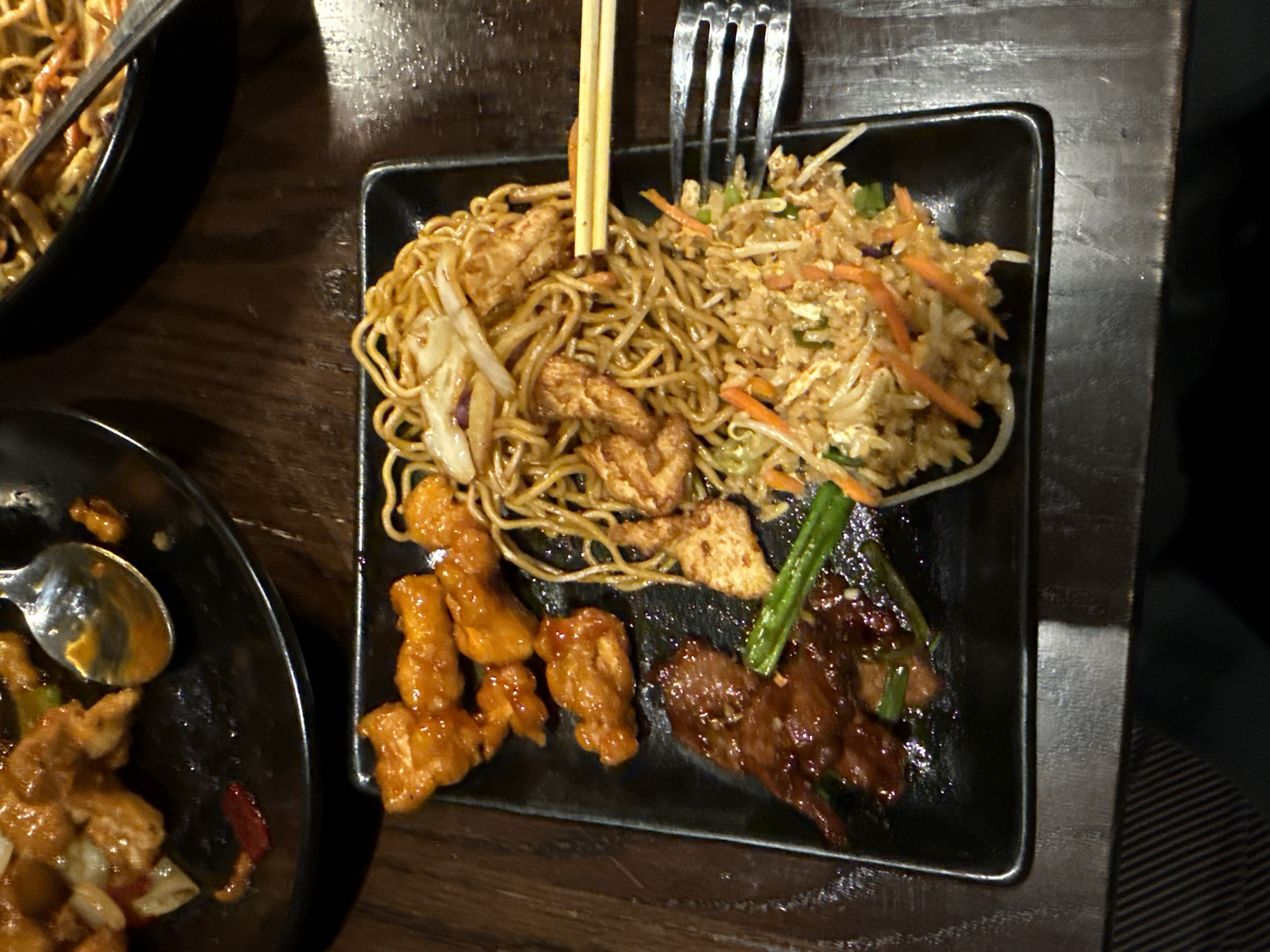 The image shows a meal served on a square black plate. The meal consists of three distinct components: noodles mixed with pieces of cooked chicken, fried rice containing carrots and green onions, and pieces of glazed chicken and beef. Utensils, including chopsticks and a fork, are visible atop the plate. Partially visible in the image are additional black dishes containing similar food items, set on a dark wooden surface. The location is the city of Chula Vista, United States of America.