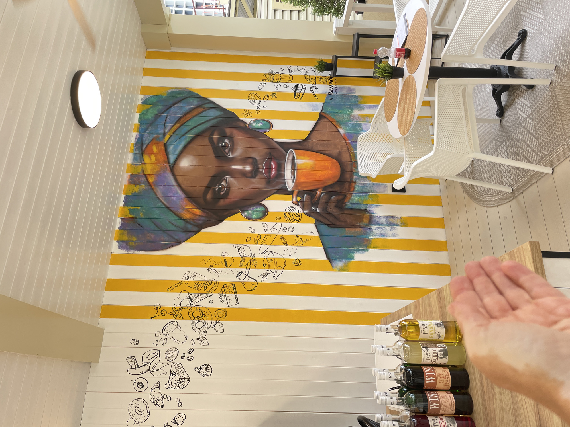 This image depicts an outdoor cafe with a vibrant mural on the wall. The mural features a woman with a colorful headscarf holding an orange cup. To the left of the mural, there are black and white drawings of various food items. The cafe is furnished with white chairs and a round white table with placemats and a bottle of water. Several bottles of syrups are arranged on a wooden counter in the foreground. The cafe appears to be located in Sochi, Russia, as indicated by the hashtag "#РусланКара", which translates to "#RuslanKara" and is a popular hashtag on social media associated with cafes in Sochi.